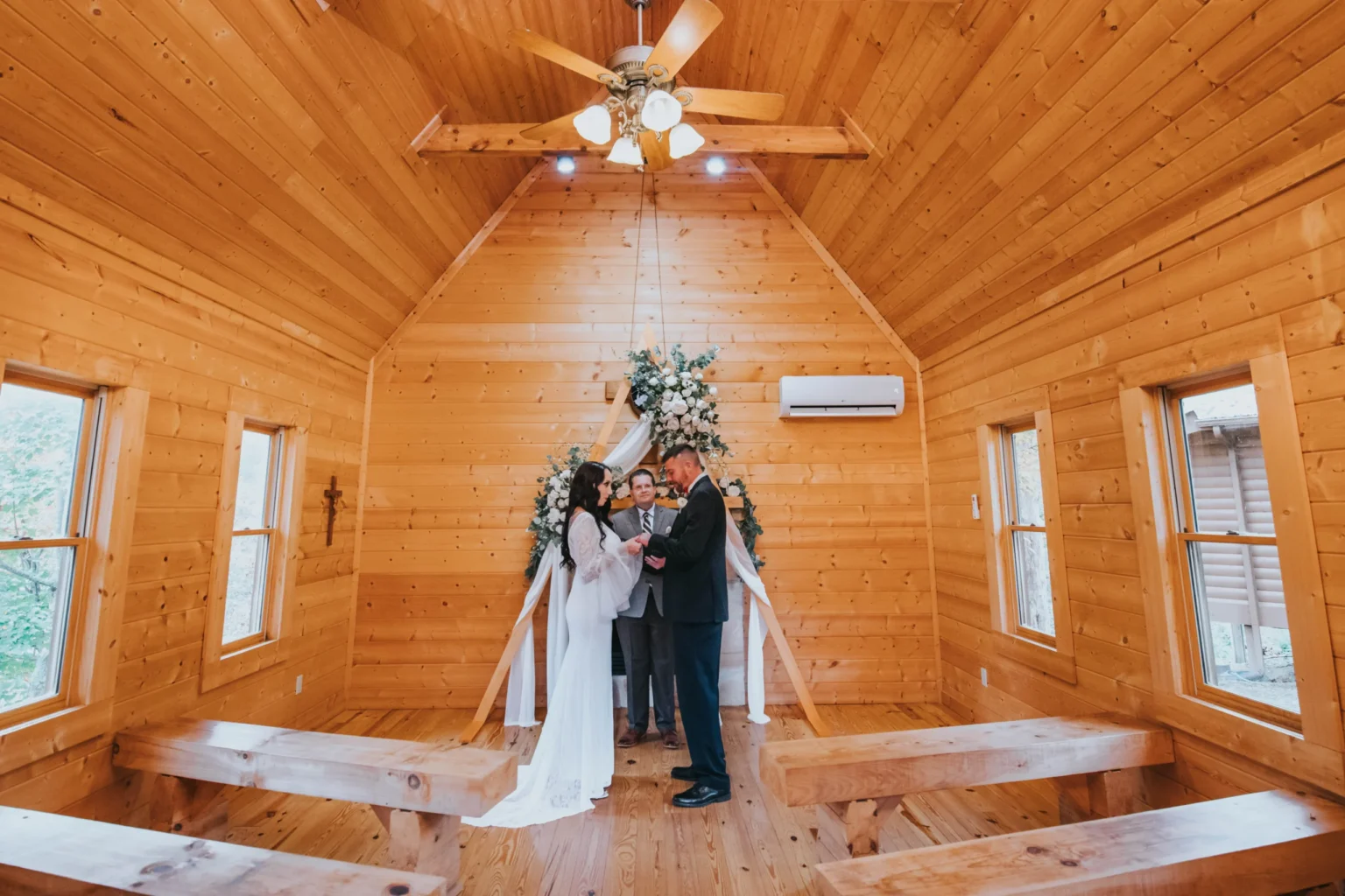 Elopement Wedding A couple stands at the altar exchanging vows in a small, wooden chapel adorned with floral arrangements. The bride wears a long white gown, and the groom is in a dark suit. A figure stands between them serving as the officiant. The chapel has wooden benches and a high ceiling with a fan. Elopements Inc