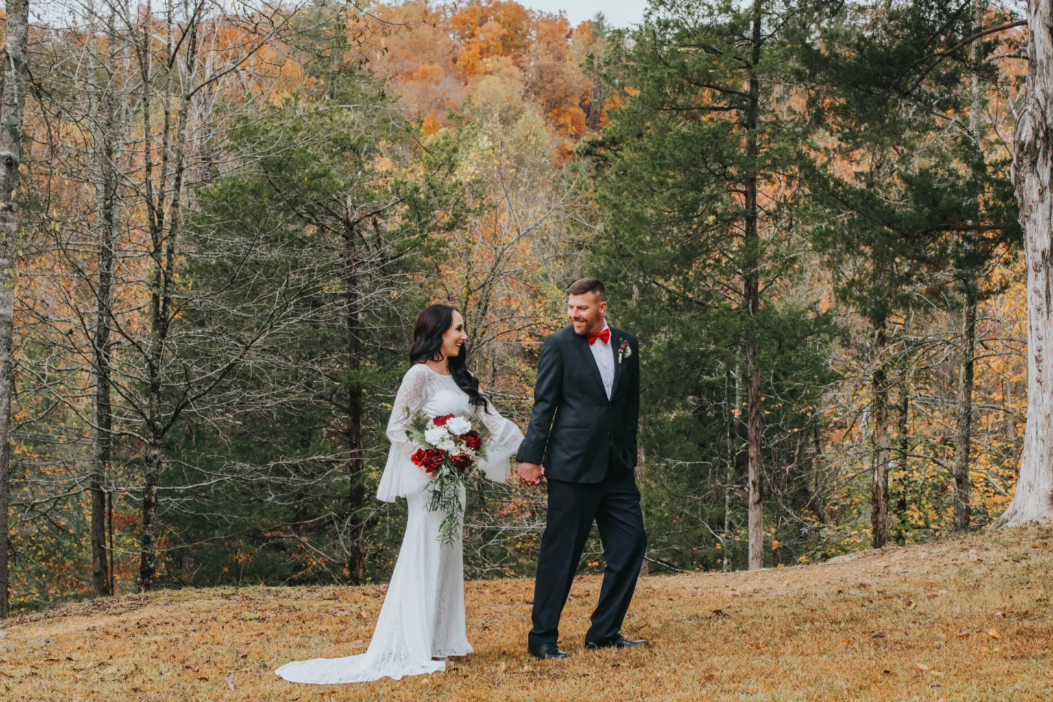 Elopement Wedding A couple is walking hand in hand outdoors among autumn trees. The woman wears a flowing white dress and holds a bouquet of red and white flowers. The man is dressed in a black suit with a red boutonniere. Both smile at each other as they walk on a grassy landscape with a backdrop of fall colors. Elopements Inc