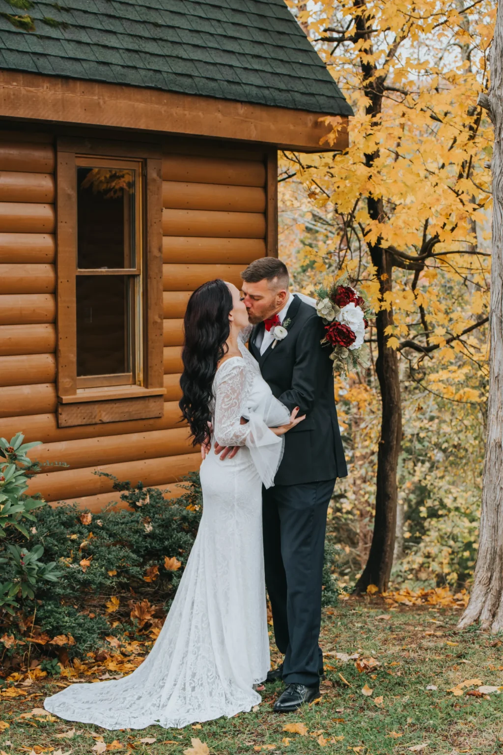 Elopement Wedding A couple stands outside a wooden cabin in an autumn setting, sharing a kiss. The bride is dressed in a white lace gown, and the groom wears a black suit with a red tie. He holds a bouquet of red and white flowers behind her back. The ground is scattered with fallen leaves, and trees with yellow foliage are visible. Elopements Inc