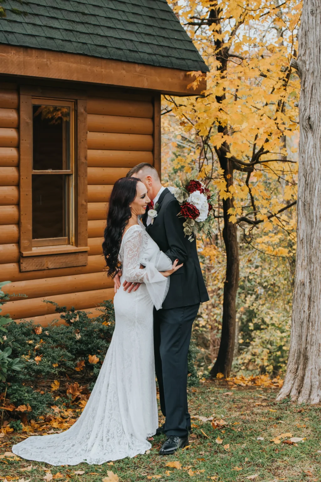 Elopement Wedding A bride and groom embrace in front of a log cabin surrounded by autumn foliage. The bride, wearing a white lace gown, holds a bouquet of red and white flowers. The groom, dressed in a black suit, kisses her forehead. The scene is calm and picturesque, with golden leaves in the background. Elopements Inc