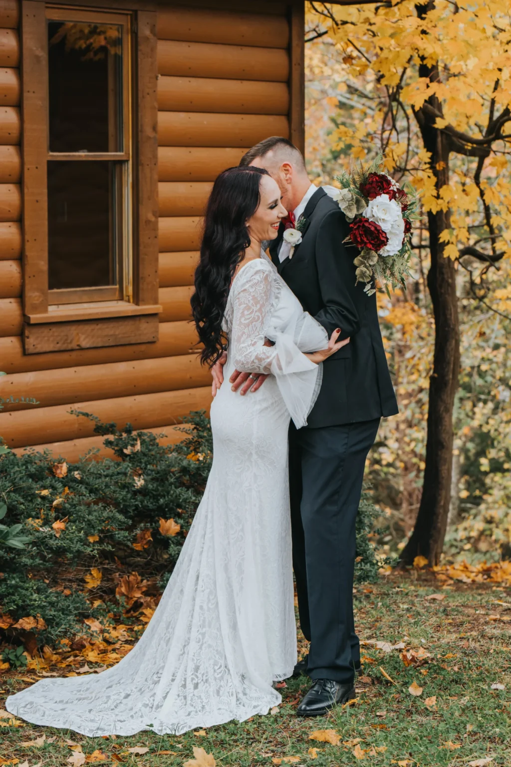 Elopement Wedding A couple stands outdoors in front of a wooden cabin, embracing. The woman is wearing a white, lacy, long-sleeved wedding dress with a train. The man, dressed in a black suit and bow tie, holds a bouquet of red and white flowers. Autumn leaves and greenery surround them. Elopements Inc