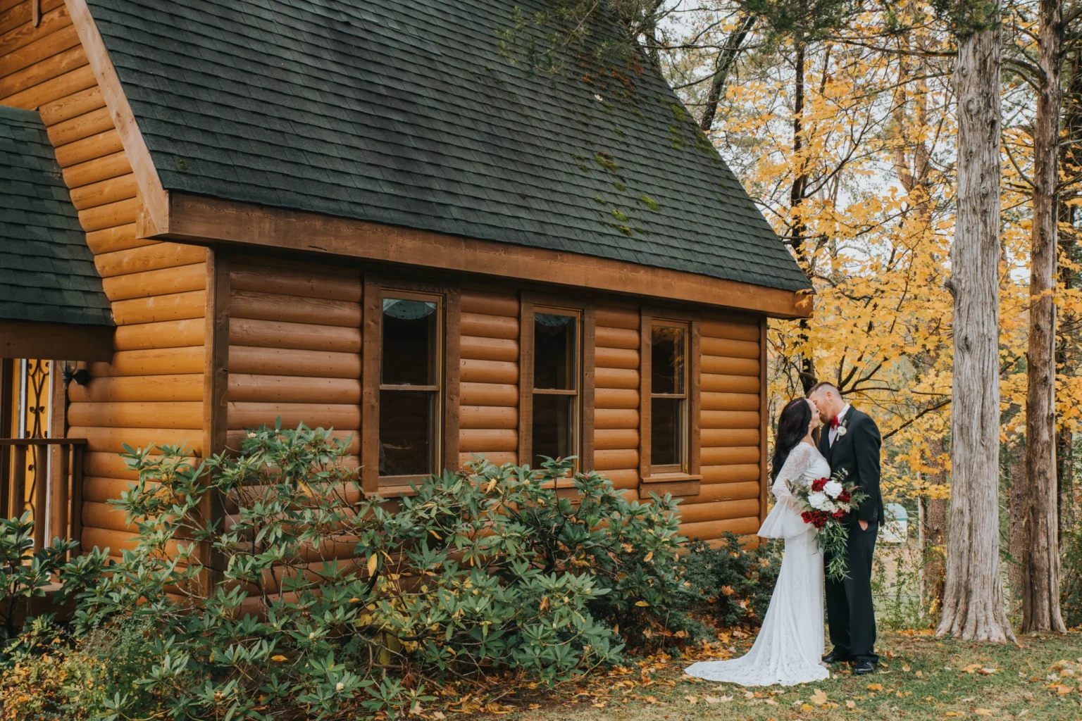 Elopement Wedding A bride and groom share a kiss beside a rustic log cabin surrounded by lush, green bushes and tall trees with vibrant yellow autumn leaves. The bride is in a white dress holding red and white flowers, while the groom is in a black suit. The setting exudes a romantic, forest ambiance. Elopements Inc