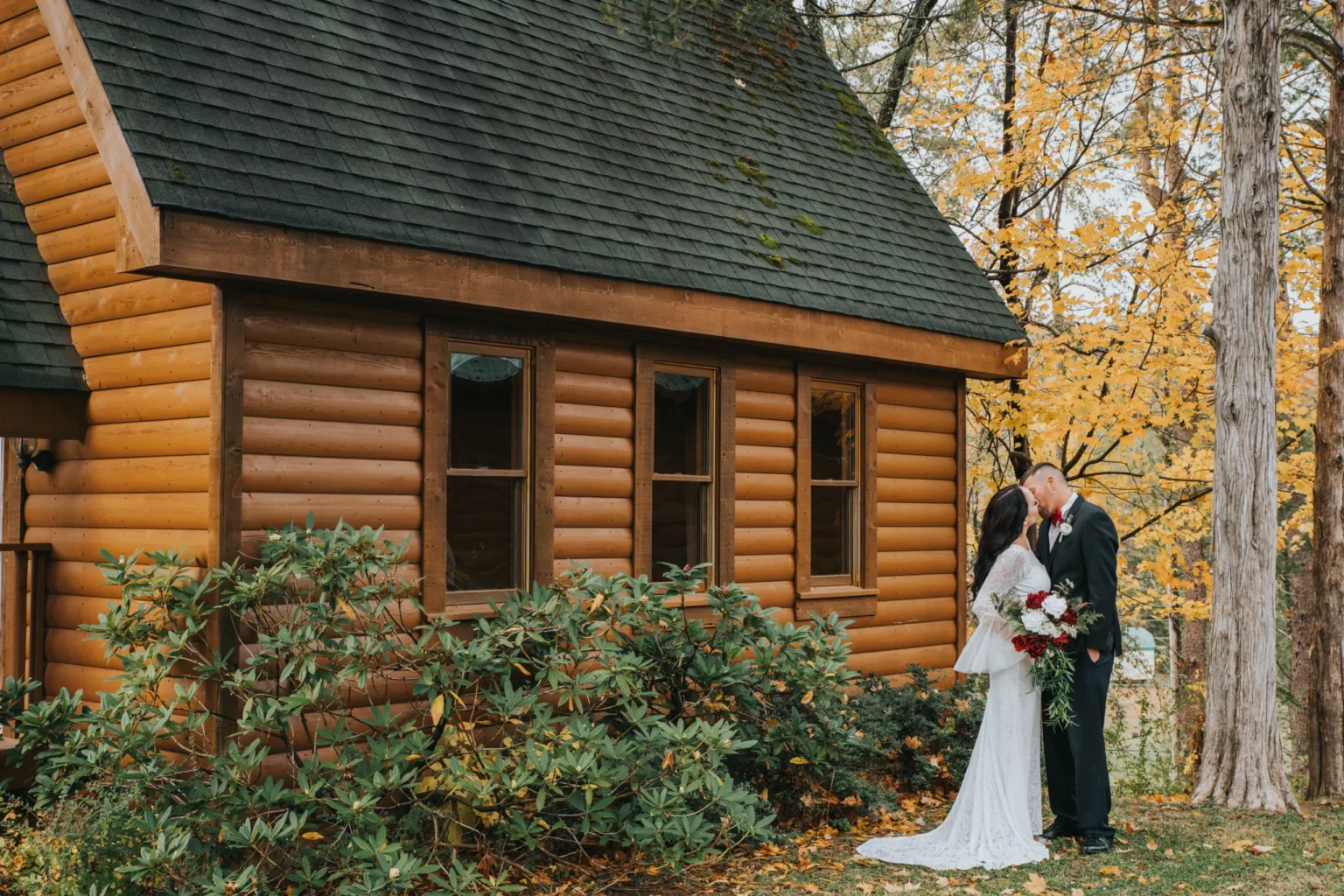 Elopement Wedding A bride in a white dress and a groom in a dark suit share a romantic kiss beside a rustic wooden cabin surrounded by lush greenery and autumn trees with golden leaves. The serene setting and vibrant fall colors add charm to the couple's special moment. Elopements Inc