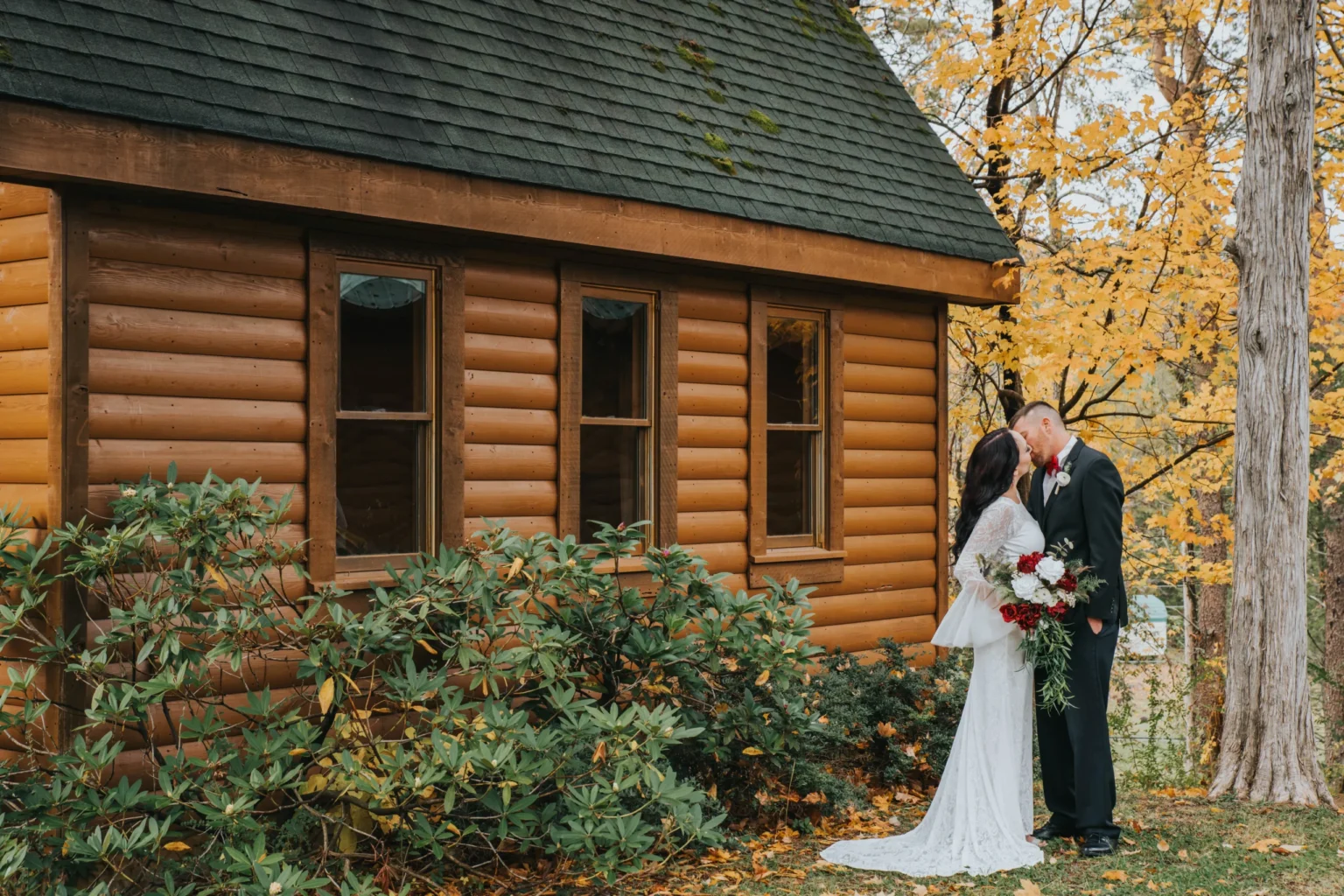 Elopement Wedding A couple in wedding attire shares a kiss beside a rustic log cabin. The bride, in a white gown, holds a bouquet of red and white flowers. The groom, in a dark suit, stands close. The backdrop features vibrant autumn foliage, creating a picturesque, romantic scene. Elopements Inc