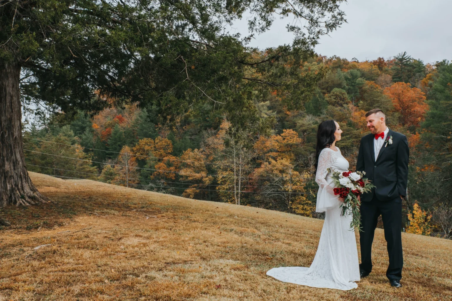 Elopement Wedding A bride and groom stand on a grassy hill surrounded by fall foliage. The bride wears a long white dress with lace sleeves and holds a bouquet of red and white flowers. The groom wearing a black suit with a red bow tie smiles at her. A large tree and forested hills are in the background. Elopements Inc