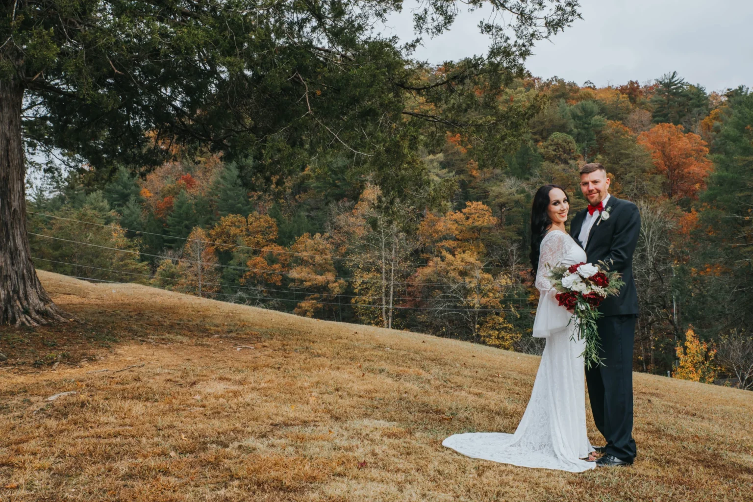 Elopement Wedding A bride in a white gown and a groom in a dark suit stand on a hillside with autumn foliage in the background. The bride holds a bouquet of red and white flowers, and they smile while embracing under a large tree with a sprawling green canopy. The ground is covered in light brown grass. Elopements Inc