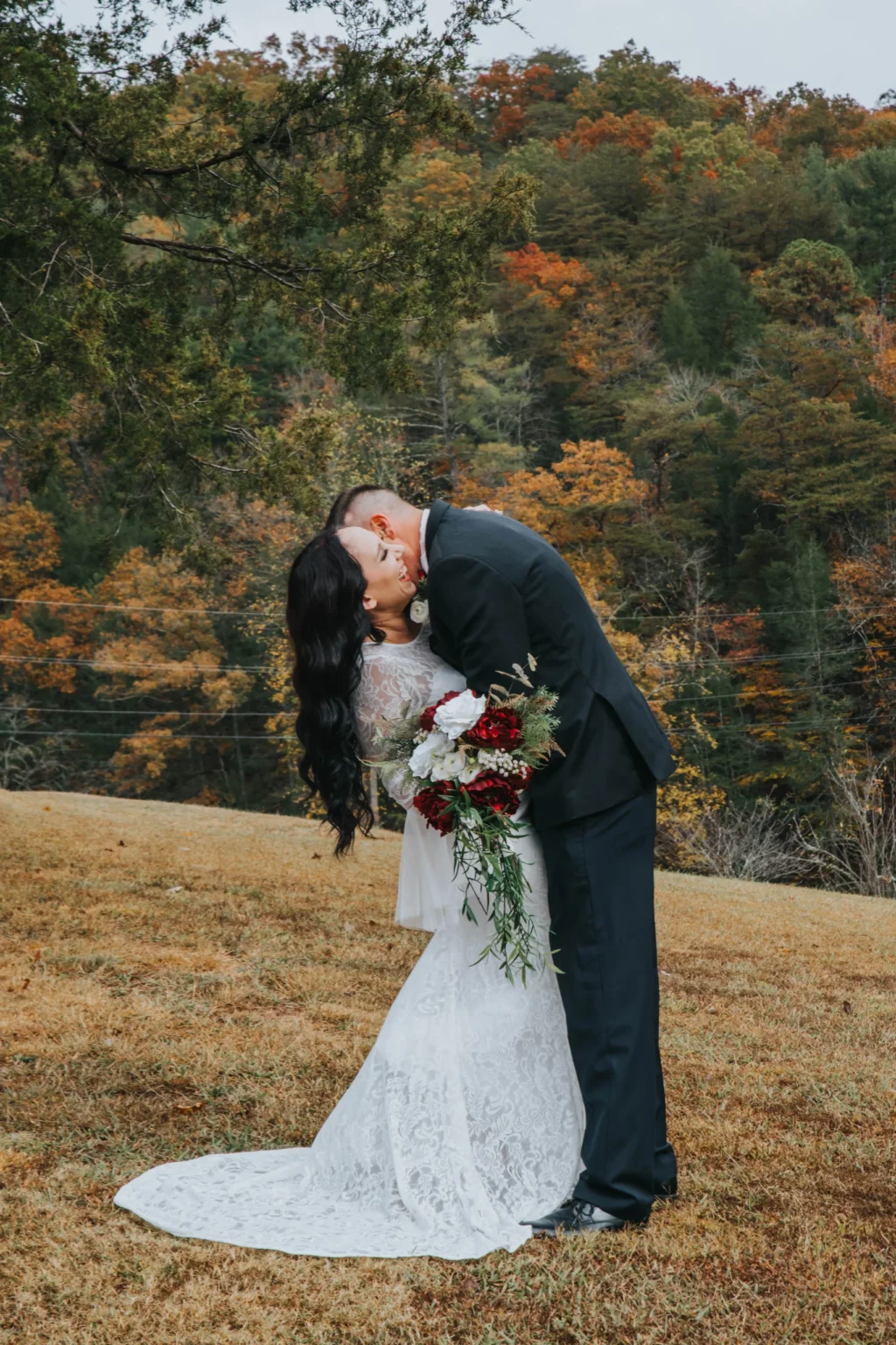 Elopement Wedding A groom in a black suit and a bride in a white lace gown share a kiss outdoors, surrounded by fall foliage. The bride holds a bouquet of red and white flowers with greenery. They stand on a grassy slope with a backdrop of colorful autumn trees. Elopements Inc