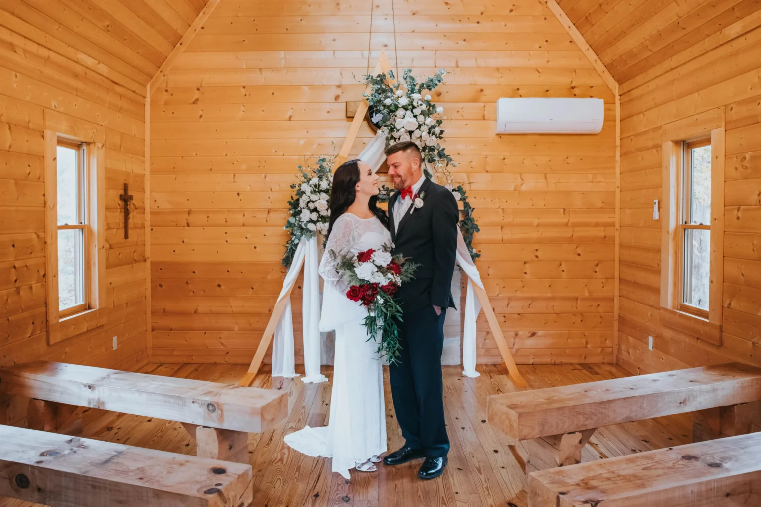 Elopement Wedding A bride and groom stand in a small wooden chapel, facing each other and smiling. The bride is holding a bouquet of red and white flowers, while the groom wears a dark suit with a boutonnière. Behind them, a floral arrangement decorates the front of the chapel. Wooden benches line the sides. Elopements Inc