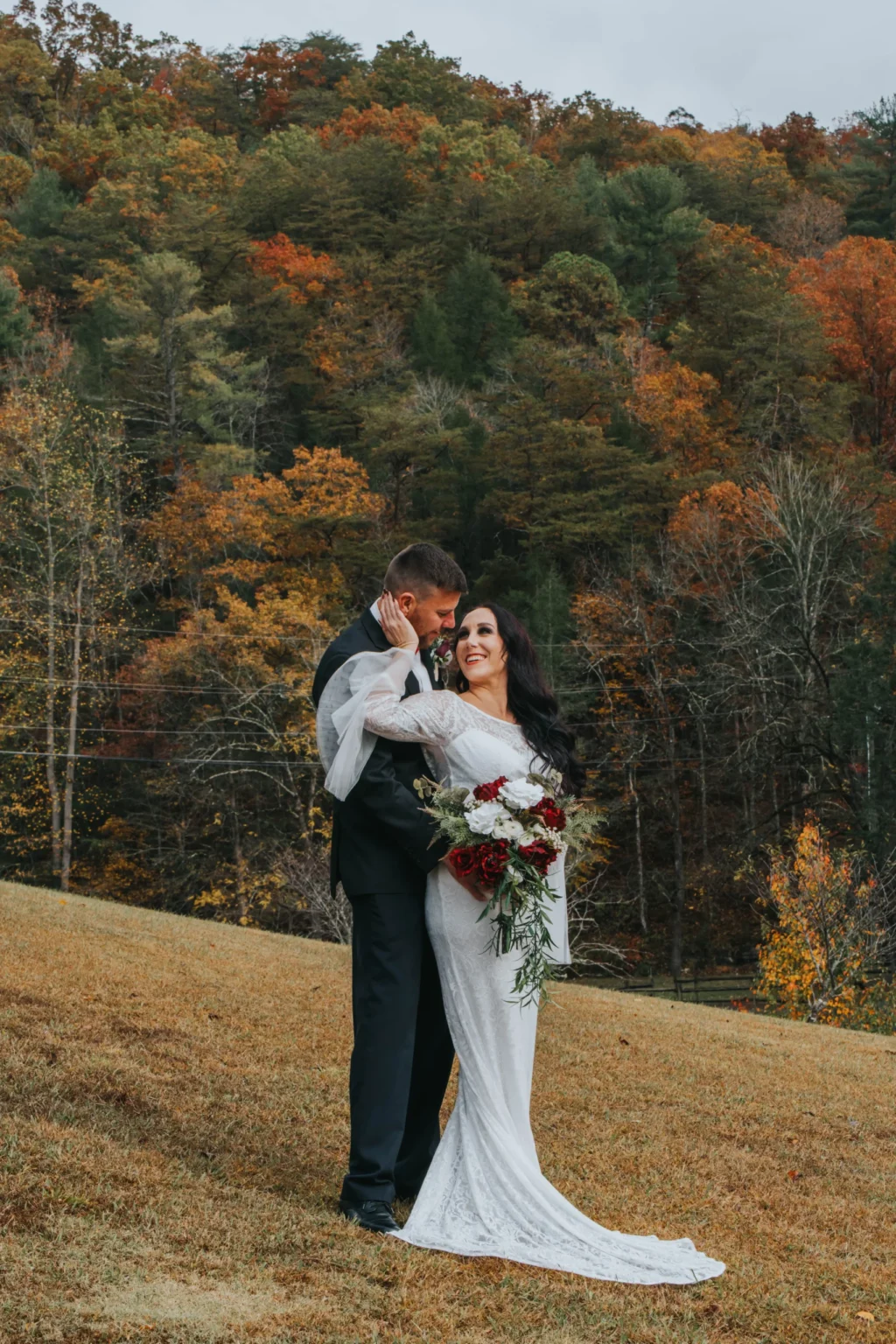 Elopement Wedding A couple stands on a grassy hill in front of a forest with autumn foliage. The groom, wearing a black suit, embraces the bride, who is in a long-sleeve white dress holding a bouquet of red and white flowers. Both are smiling and looking at each other lovingly. The sky above is overcast. Elopements Inc