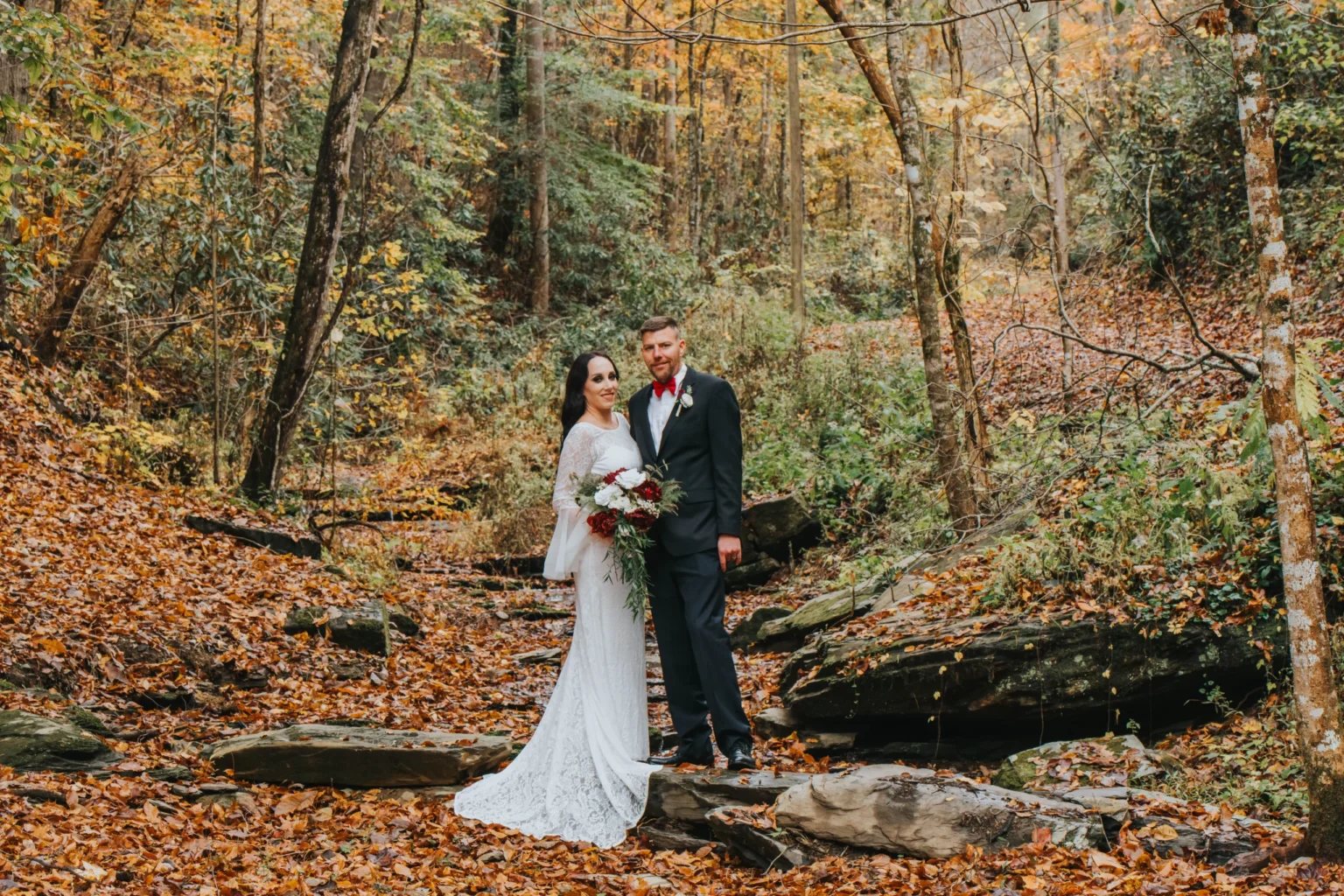 Elopement Wedding A bride in a white dress and groom in a dark suit stand together in a forest with autumn foliage. The bride holds a bouquet of red and white flowers. They pose on large rocks near a small stream, surrounded by trees with colorful leaves, creating a serene and picturesque backdrop. Elopements Inc