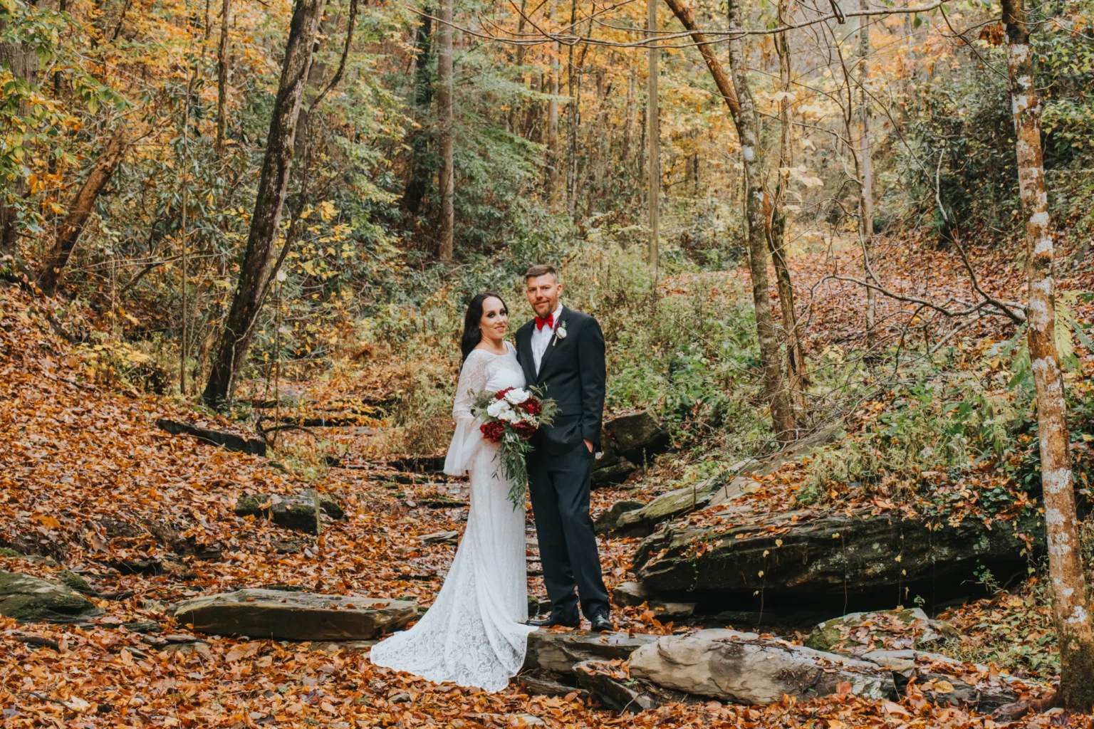 Elopement Wedding A bride in a white lace gown and the groom in a black suit with a red tie stand together on rocks in a forest blanketed with autumn leaves. The bride holds a bouquet of red and white flowers. Tall trees with fall foliage surround the couple as they pose in a serene woodland setting. Elopements Inc