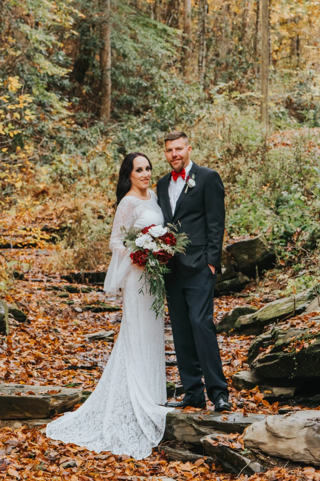 Elopement Wedding A couple stands outdoors in a forest during autumn. The bride is wearing a long-sleeved white lace wedding dress and holding a bouquet of red and white flowers. The groom is wearing a black suit with a red bow tie and boutonniere. Fallen leaves in shades of orange and yellow cover the ground. Elopements Inc