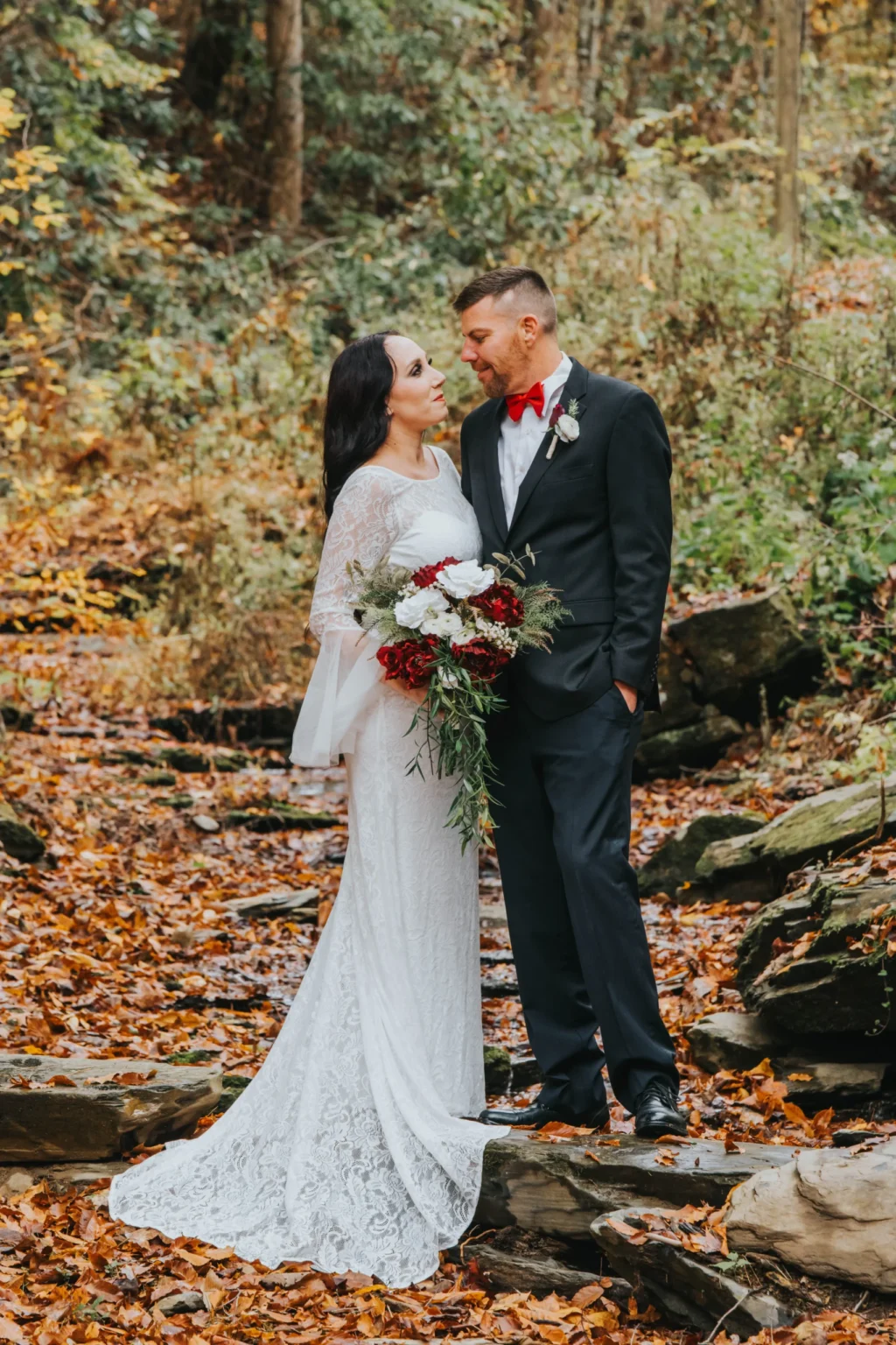 Elopement Wedding A bride and groom stand together in a wooded area with fallen autumn leaves. The bride wears a long white lace dress and holds a bouquet of red and white flowers. The groom is dressed in a dark suit with a red bow tie and boutonniere. They are gazing lovingly at each other. Elopements Inc