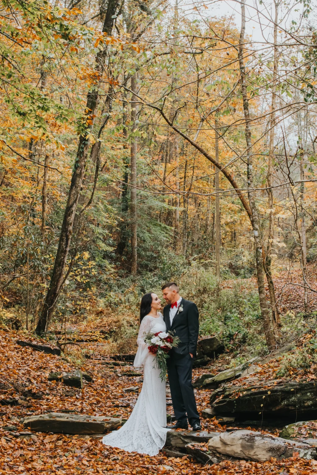 Elopement Wedding A couple stands in a forest filled with autumn leaves. The bride, in a white lace gown, holds a bouquet of red and white flowers and gazes up at the groom, who wears a dark suit with a red bow tie. They stand among rocks and fallen leaves, surrounded by trees with yellow and orange foliage. Elopements Inc