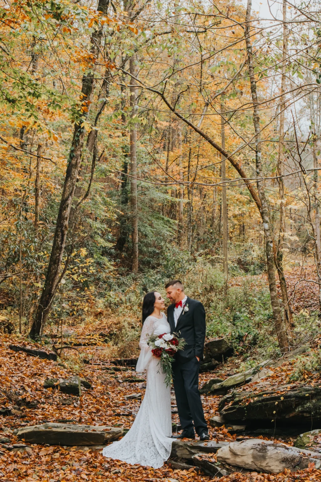 Elopement Wedding A bride in a long white dress holding a bouquet and a groom in a black suit with a red bow tie share an intimate moment in an autumn forest. They are surrounded by fallen leaves, tall trees, and a rocky terrain, creating a serene and romantic atmosphere. Elopements Inc