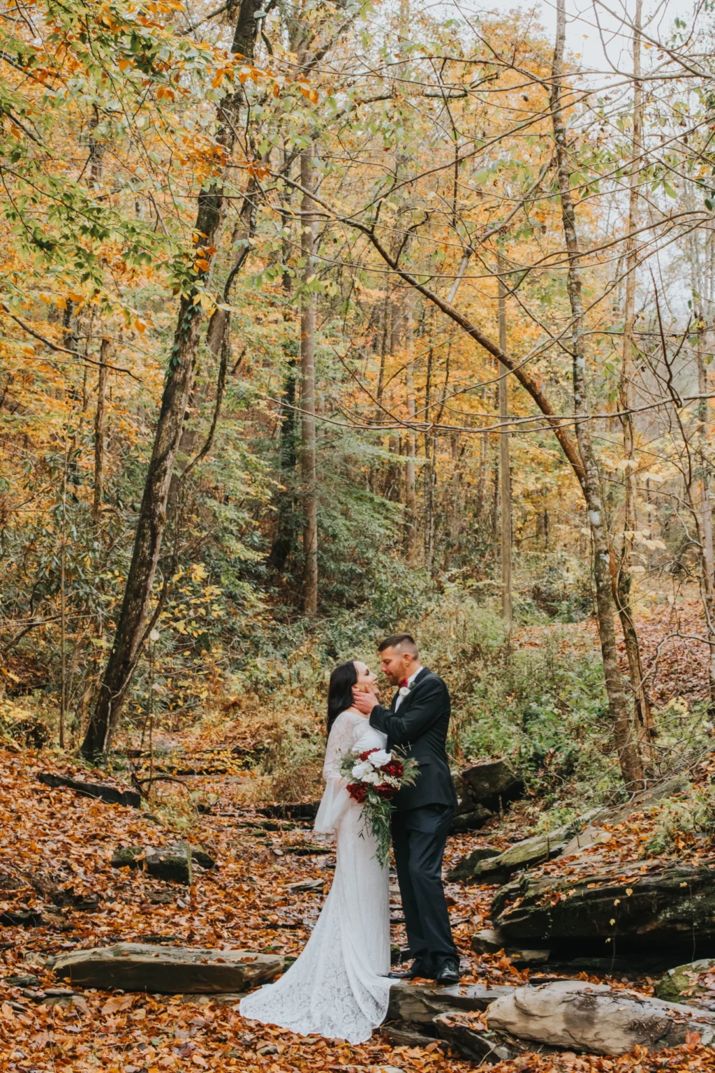 Elopement Wedding A bride and groom stand in an autumnal forest, surrounded by trees with yellow and orange leaves. The bride, in a white lace gown, holds a bouquet of red and white flowers. The groom, in a black suit, embraces her. They stand on rocks amidst fallen leaves, with a serene, natural backdrop. Elopements Inc