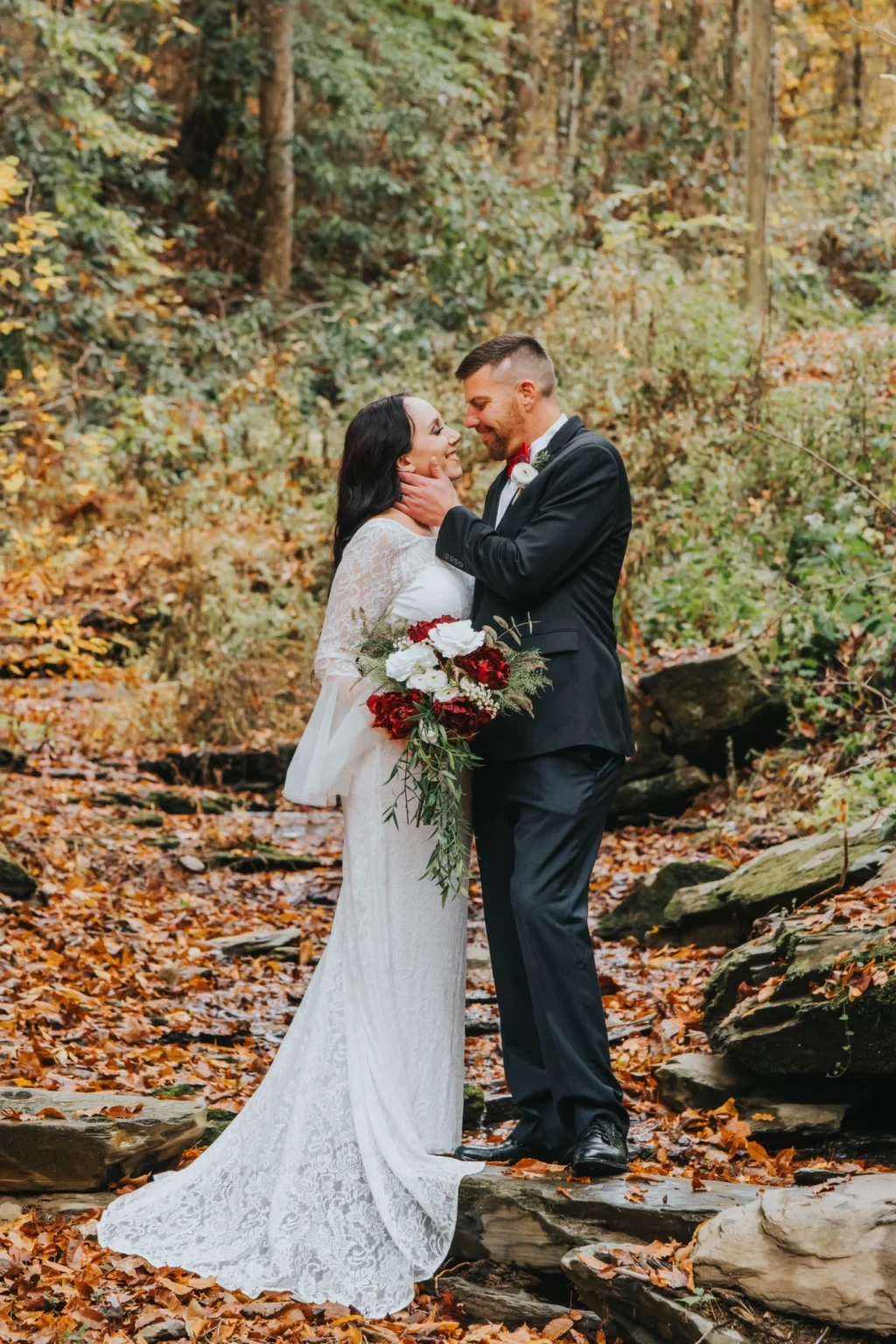 Elopement Wedding A couple embraces in a wooded area during autumn. The bride wears a long white lace gown and the groom is in a black suit with a red bow tie. She holds a bouquet of red and white flowers. The ground is covered in fallen leaves and there is greenery in the background. Elopements Inc