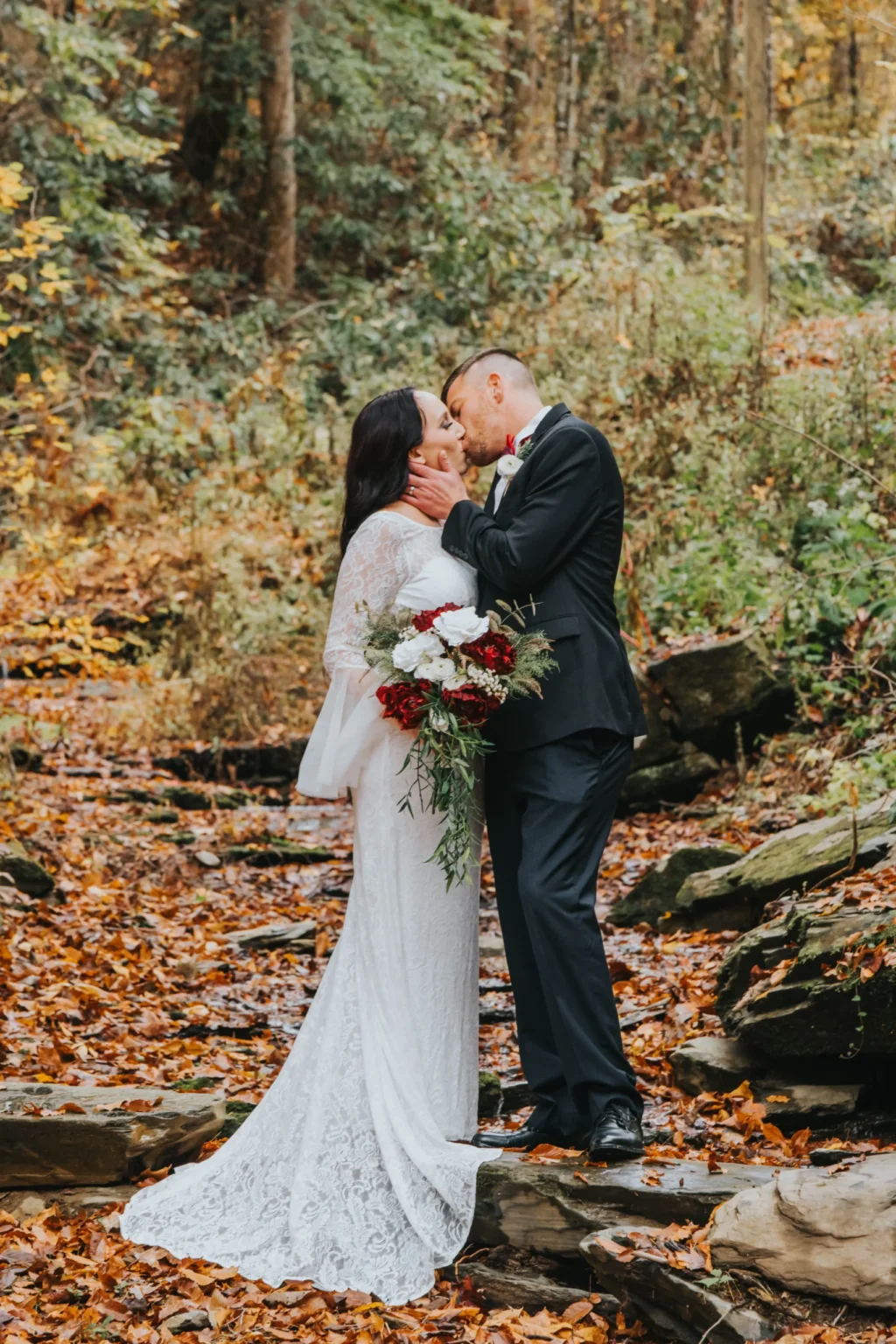 Elopement Wedding A couple in wedding attire shares a kiss in a forest setting with autumn leaves covering the ground. The bride wears a long-sleeved white lace gown and holds a bouquet of red and white flowers. The groom is in a black suit with a boutonnière, standing on stone steps surrounded by lush greenery and fallen leaves. Elopements Inc