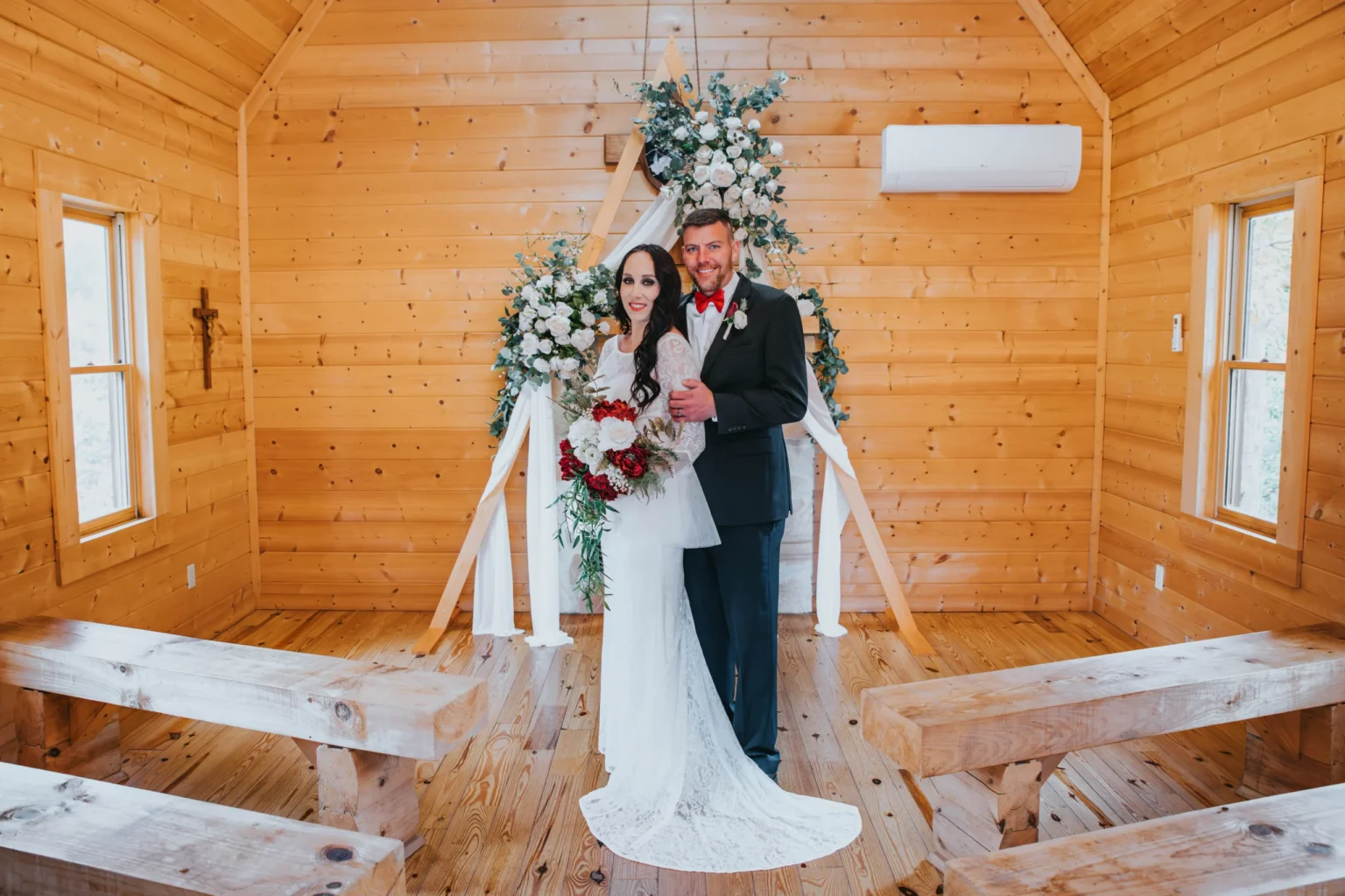 Elopement Wedding A bride in a white gown and a groom in a black suit stand arm in arm in a wooden chapel. The bride holds a bouquet of red and white flowers. Behind them is a floral arrangement with white flowers and greenery. The chapel has wooden walls, benches, and windows, with light streaming in. Elopements Inc