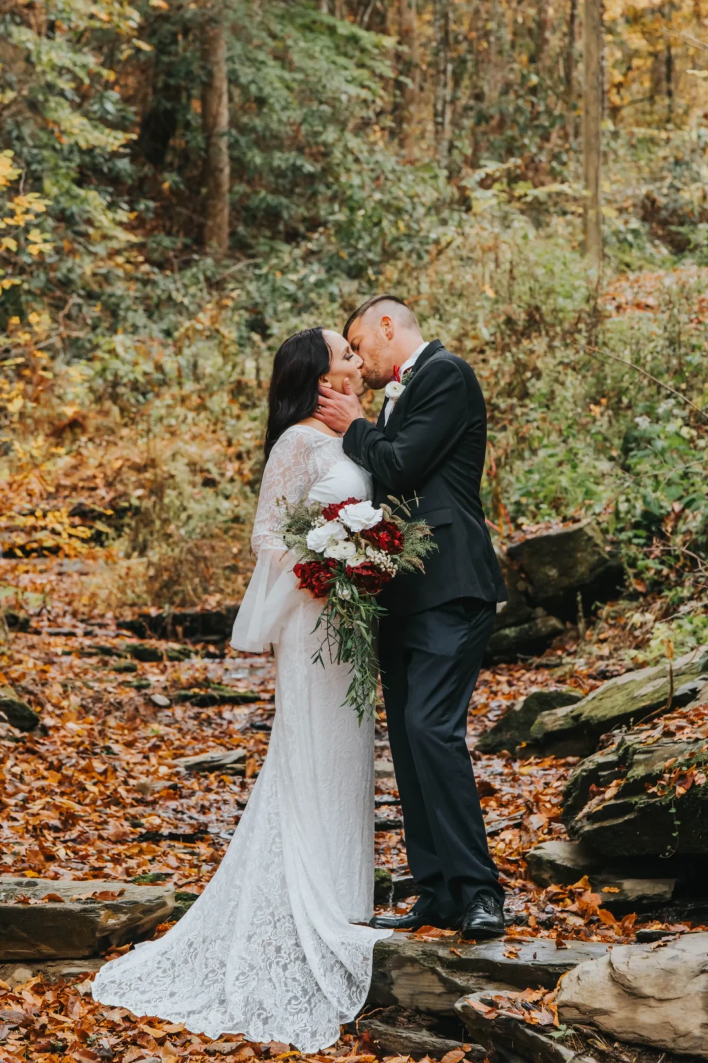 Elopement Wedding A couple kisses in a wooded area with autumn foliage. The woman, in a long white lace dress, holds a bouquet of dark red and white flowers. The man, in a black suit, gently cradles her face. They stand on a path covered in fallen leaves, with greenery and trees surrounding them. Elopements Inc