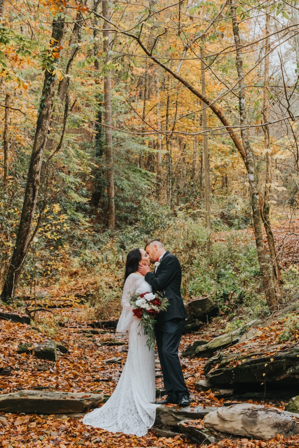 Elopement Wedding A bride in a white lace dress and a groom in a dark suit kiss while standing on a stone in a forest. They are surrounded by autumnal trees with colorful leaves. The bride holds a bouquet of red and white flowers, and fallen leaves cover the ground. Elopements Inc