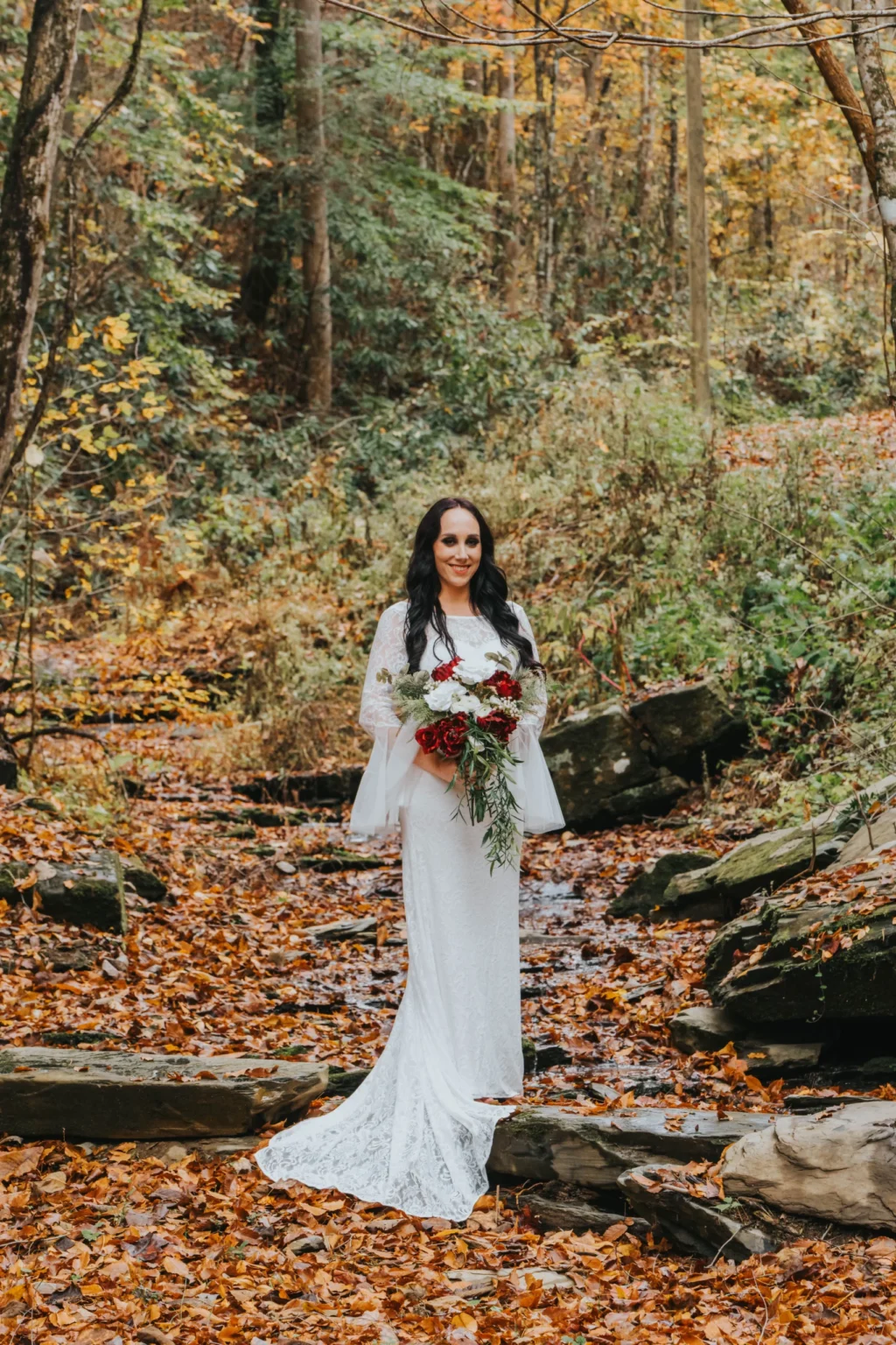 Elopement Wedding A woman stands on a rock in a forest, wearing a white lace wedding dress with long sleeves. She holds a bouquet of red and white flowers with greenery. The forest floor is covered in fallen autumn leaves, and the background is filled with trees and lush foliage. Elopements Inc