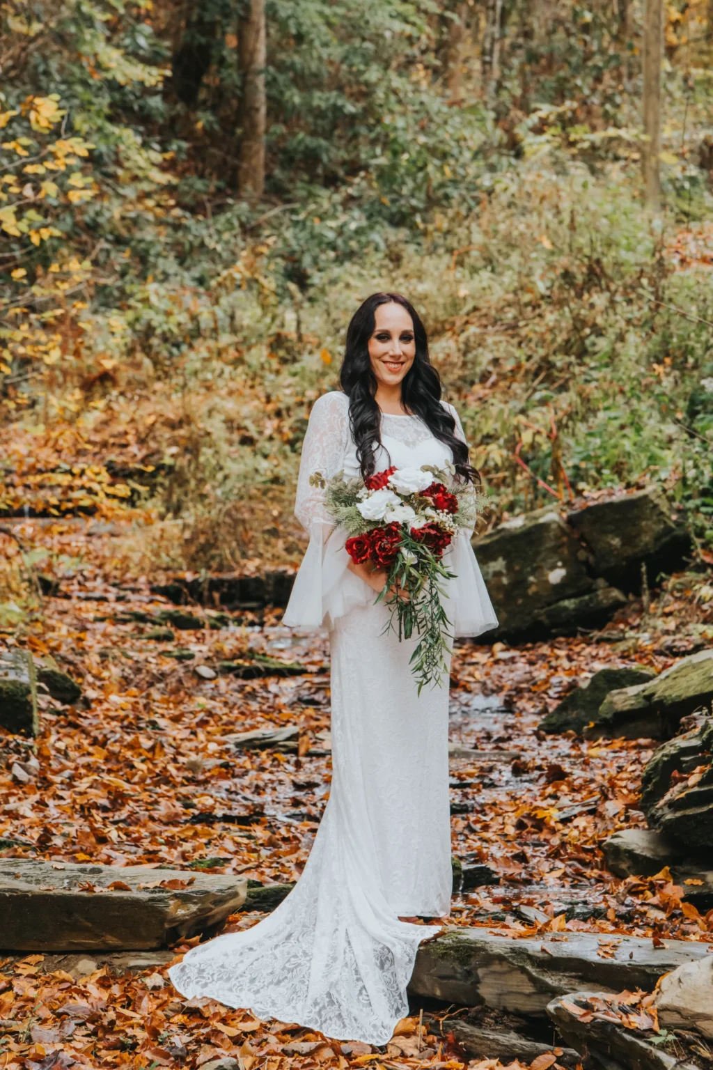 Elopement Wedding A woman stands in a forest in autumn, surrounded by fallen leaves. She is wearing a long, white lace wedding dress and holding a bouquet of red, white, and green flowers. The background features greenery and trees, creating a rustic setting. She looks at the camera with a serene expression. Elopements Inc