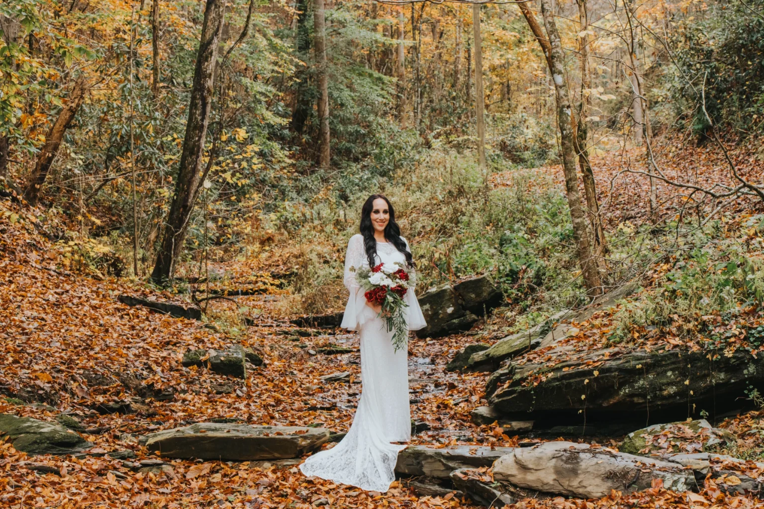 Elopement Wedding A bride in a white lace gown stands among rocks and fallen leaves in a forest during autumn. She holds a large bouquet of white and red flowers with greenery. The trees surrounding her display vibrant yellow, orange, and green leaves, creating a picturesque backdrop. Elopements Inc