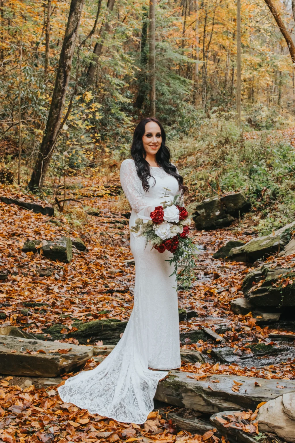 Elopement Wedding A woman in a white lace wedding dress stands on a rock in a forest filled with autumn leaves. She has long, dark hair and is holding a bouquet of red and white flowers with greenery. The background includes trees with fall-colored foliage and a forest floor covered in fallen leaves. Elopements Inc