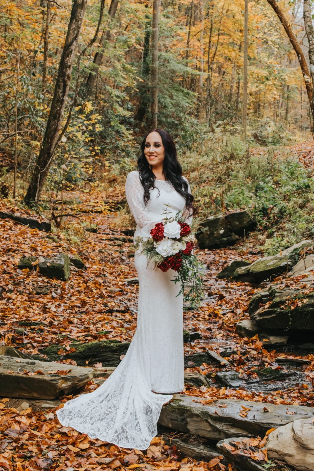 Elopement Wedding A bride stands in a forest during autumn, surrounded by colorful fallen leaves and trees with orange and yellow foliage. She wears a white lace wedding dress and holds a bouquet of red and white flowers with greenery. The scene is serene and picturesque, with rocks and a small stream nearby. Elopements Inc