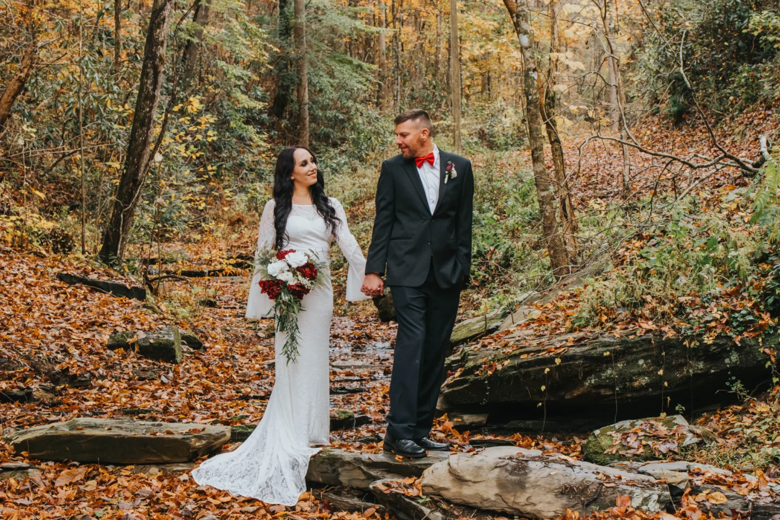 Elopement Wedding A newlywed couple walks hand-in-hand through a serene forest with autumn foliage. The bride wears a long white lace gown and holds a bouquet of red and white flowers. The groom is dressed in a black suit with a red bow tie and boutonnière. They smile at each other, surrounded by nature. Elopements Inc