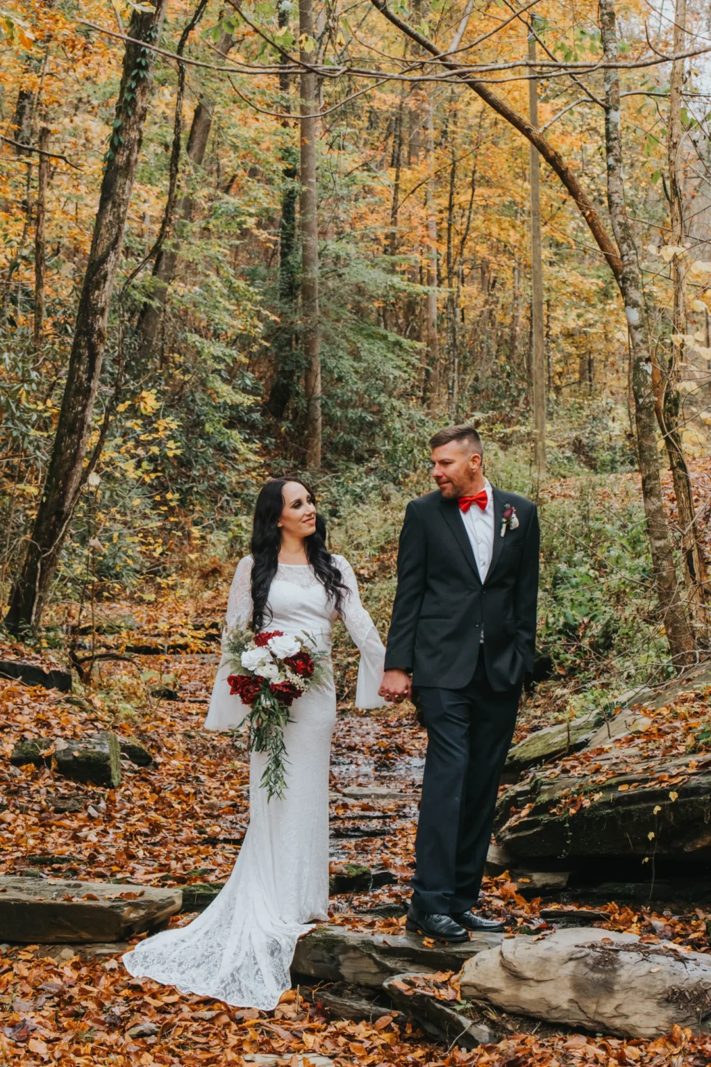 Elopement Wedding A couple stands in a woodland area with autumn leaves scattered on the ground. The bride, in a long-sleeved white gown and holding a floral bouquet, looks at the groom. The groom, dressed in a black suit with a red bow tie and boutonniere, gazes back, holding her hand. Trees with fall foliage surround them. Elopements Inc