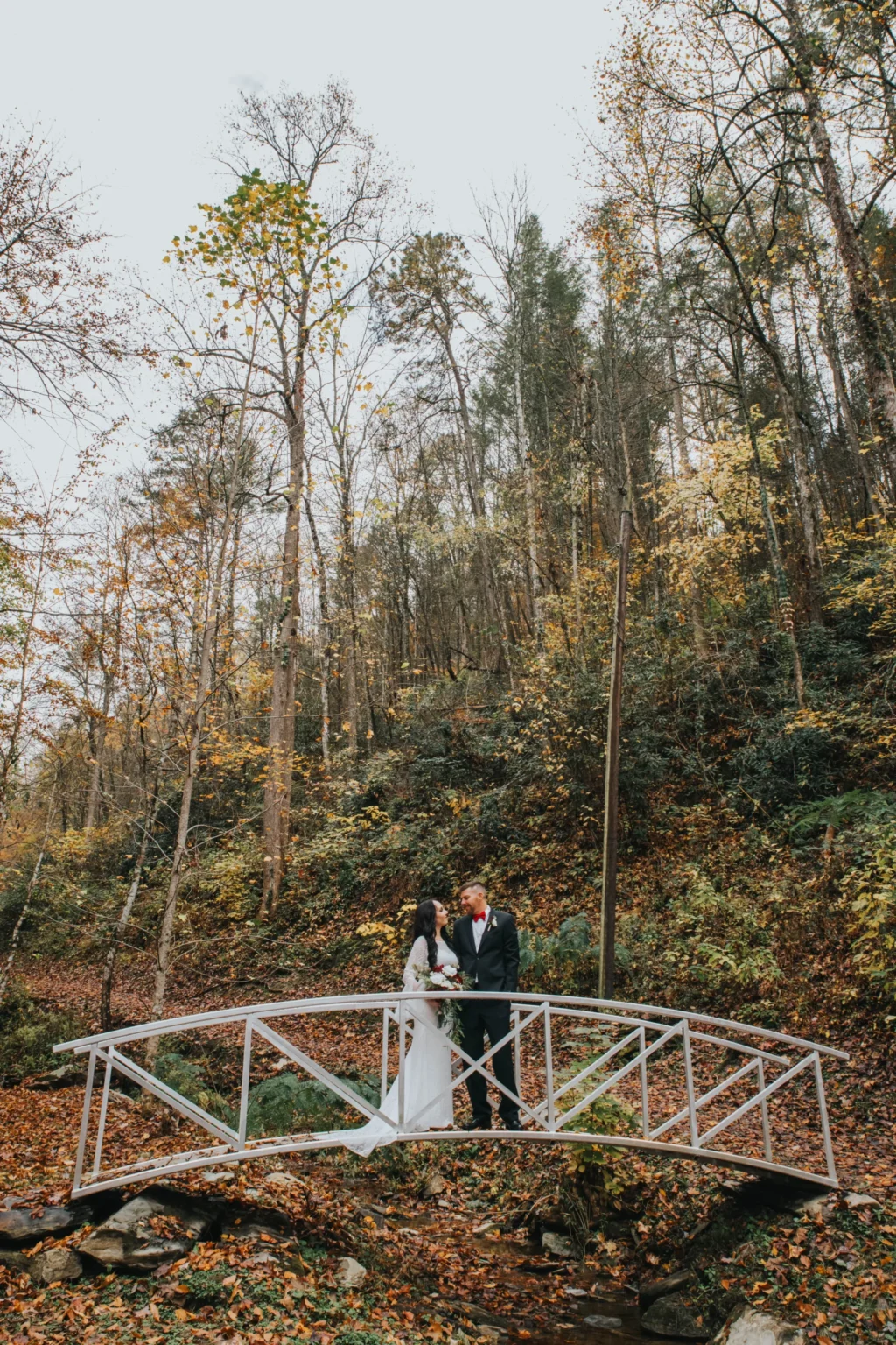 Elopement Wedding A bride and groom stand on a small metal bridge in a forest setting. The bride wears a long white dress, and the groom is in a dark suit. The surrounding trees, shedding autumn leaves, create a natural, scenic backdrop. The couple appears to be sharing a tender moment. Elopements Inc