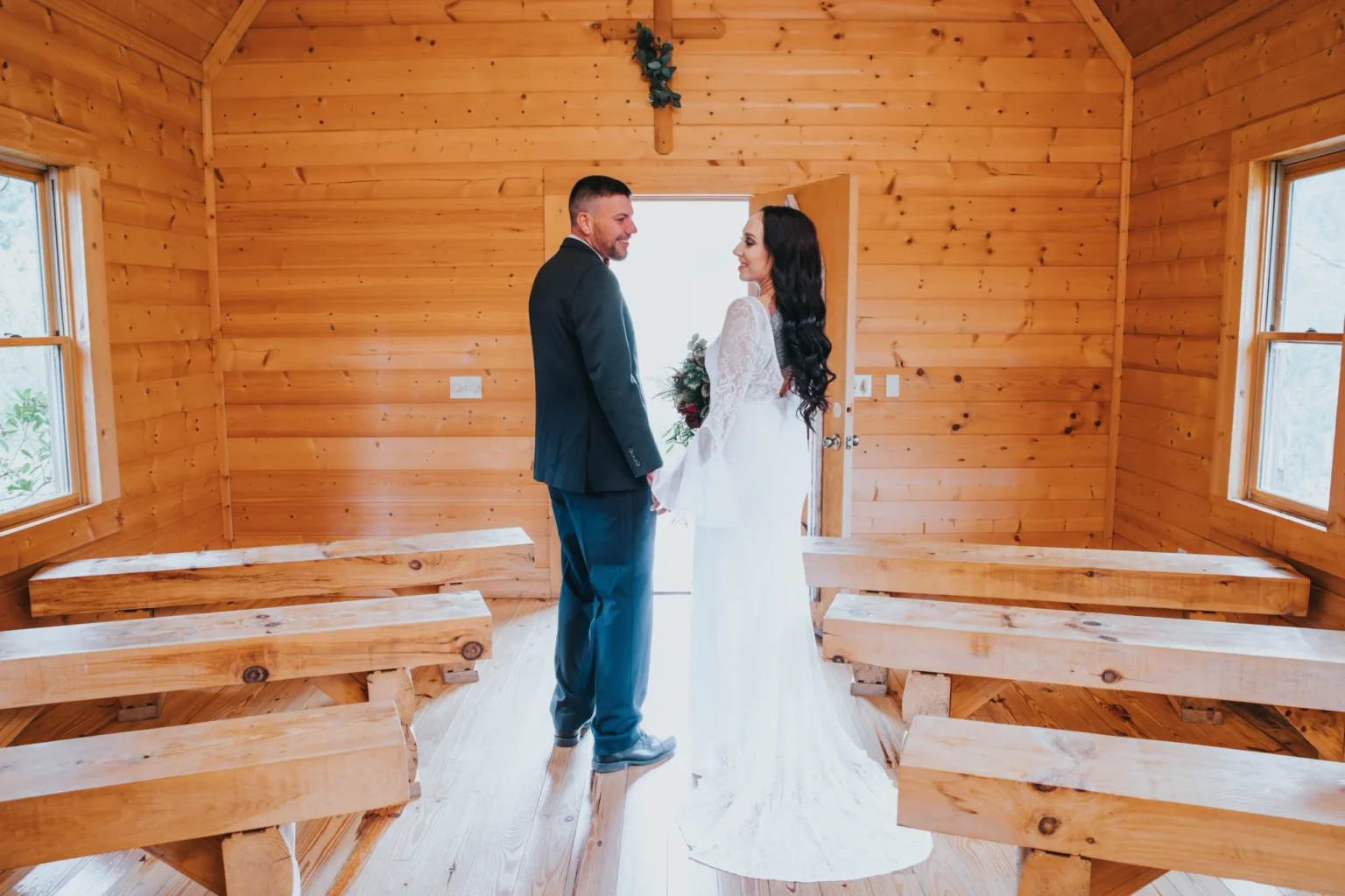 Elopement Wedding A couple stands at the doorway of a rustic wooden chapel, holding hands and smiling at each other. The groom is in a dark suit, and the bride wears a long white dress. The chapel features wooden benches and walls, with natural light coming through the windows. A small decorative wreath hangs above the door. Elopements Inc