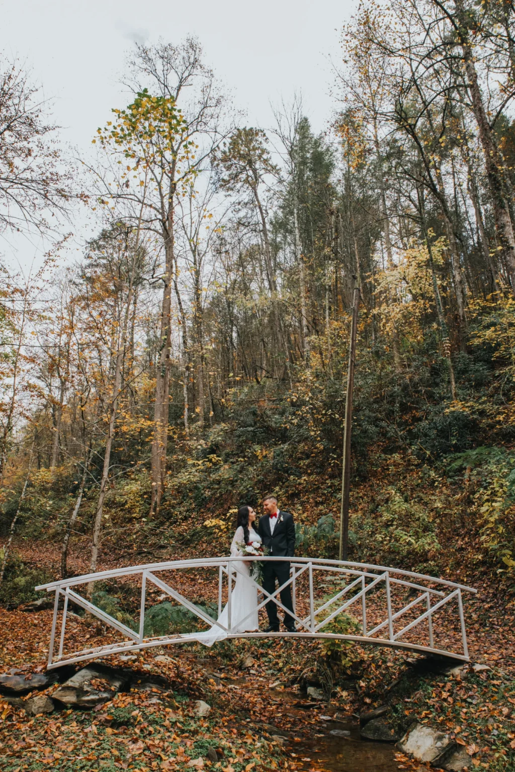 Elopement Wedding A bride and groom stand on a small white bridge in a forest. The bride wears a white dress, and the groom is in a dark suit. Surrounding them are tall trees with autumn foliage. The ground is covered with fallen leaves, creating a picturesque, romantic scene. Elopements Inc