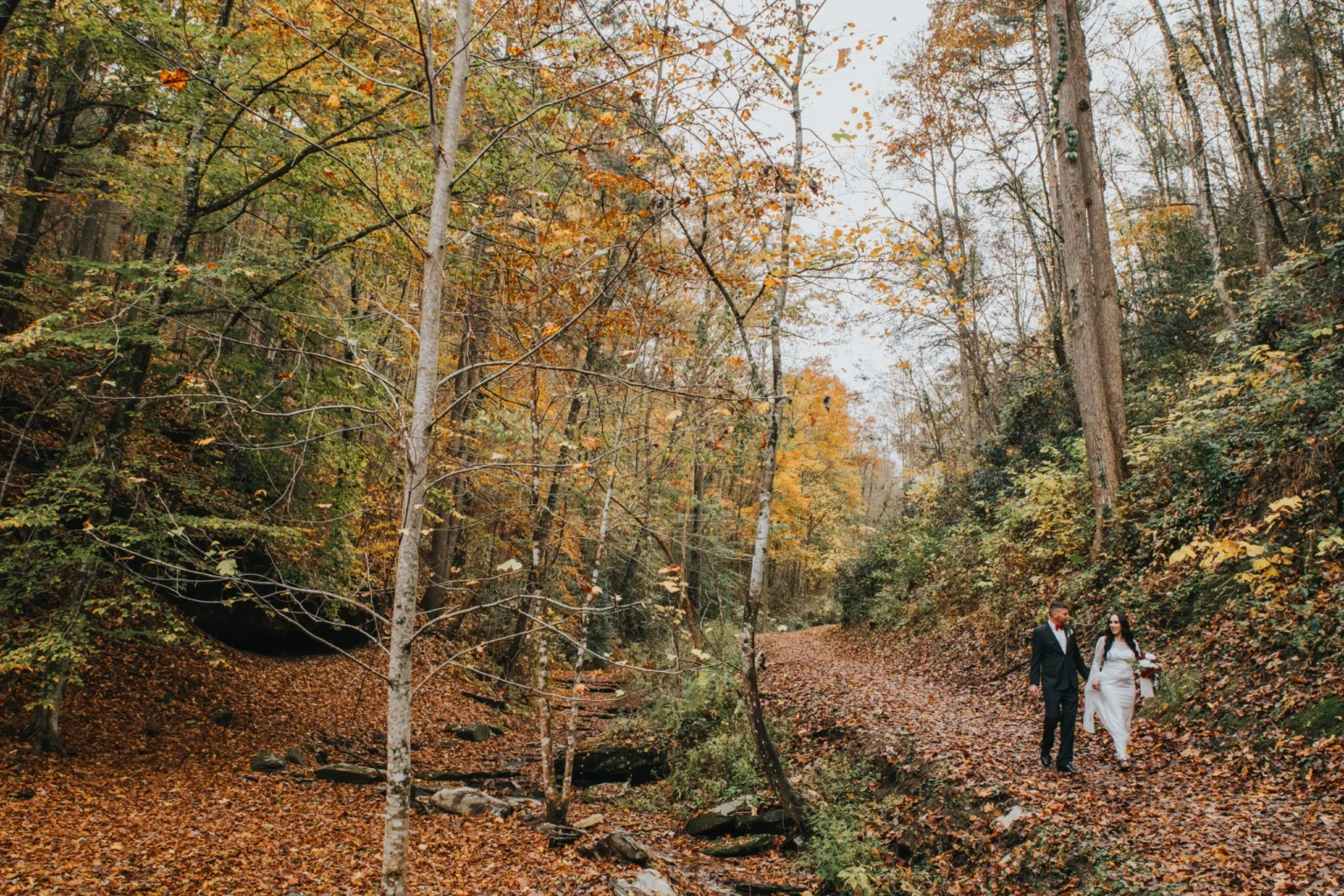 Elopement Wedding A couple dressed in formal wedding attire, with the bride in white and the groom in a dark suit, walk hand-in-hand through a forested path covered in autumn leaves. Around them, trees in hues of green, yellow, and orange create a picturesque, serene fall landscape. Elopements Inc