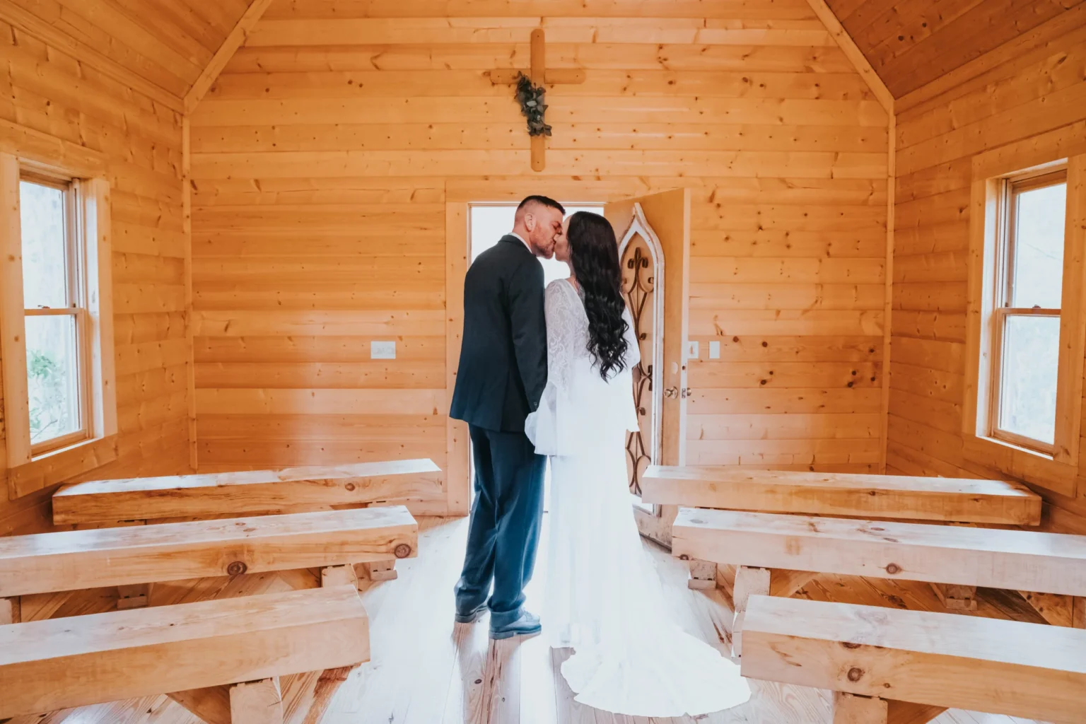 Elopement Wedding A couple in wedding attire stands at the front of a small wooden chapel, holding hands and sharing a kiss. The chapel has light wooden walls, a vaulted ceiling, and bench-style pews. A closed door with a decorative design is in the background, and sunlight streams through two windows on either side. Elopements Inc