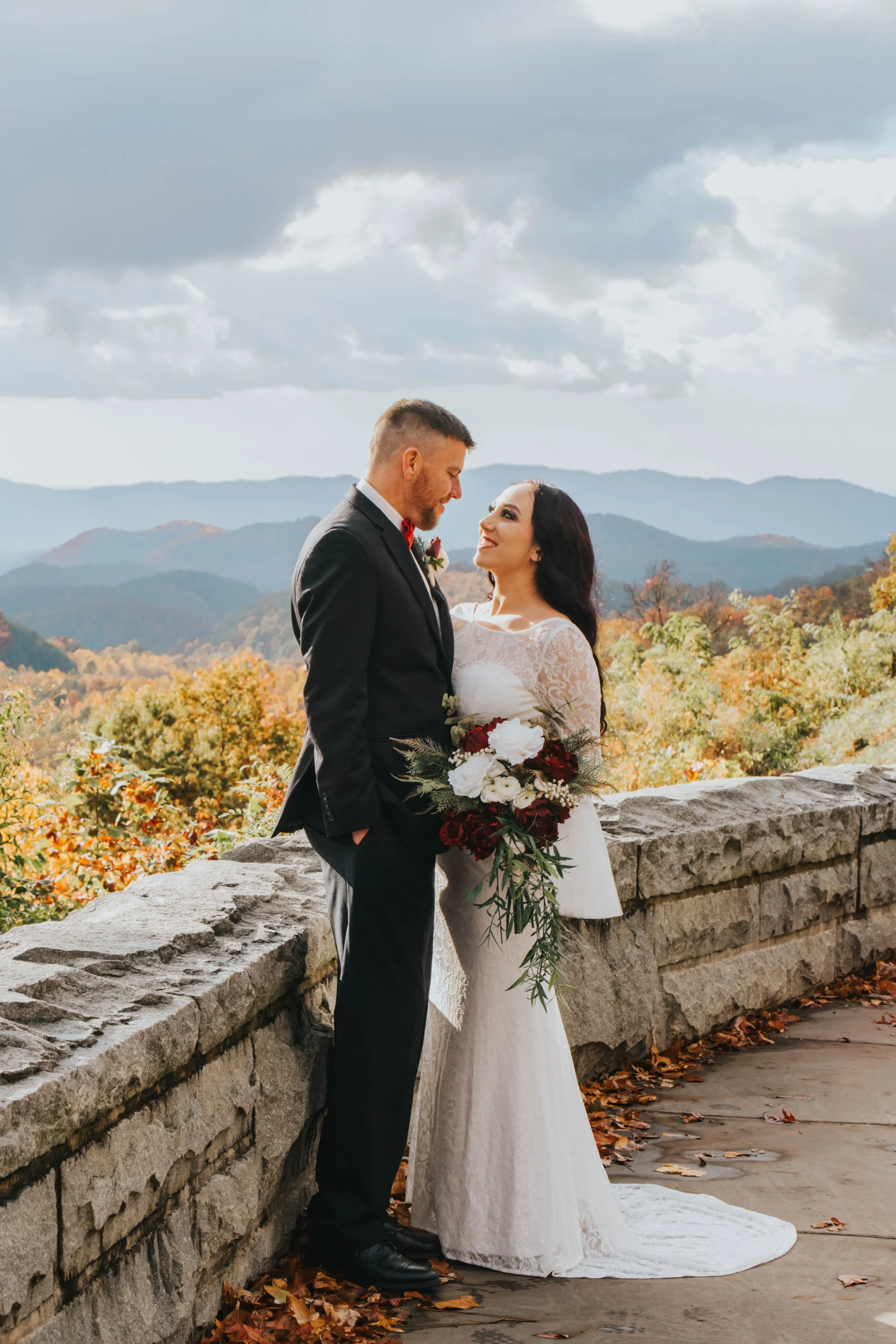 Elopement Wedding A bride and groom stand intimately, gazing at each other, on a stone pathway with a scenic mountain view in the background. The bride wears a long, white lace gown and holds a bouquet of flowers. The groom is in a black suit with a red boutonniere. The sky is partly cloudy with vibrant fall foliage. Elopements Inc