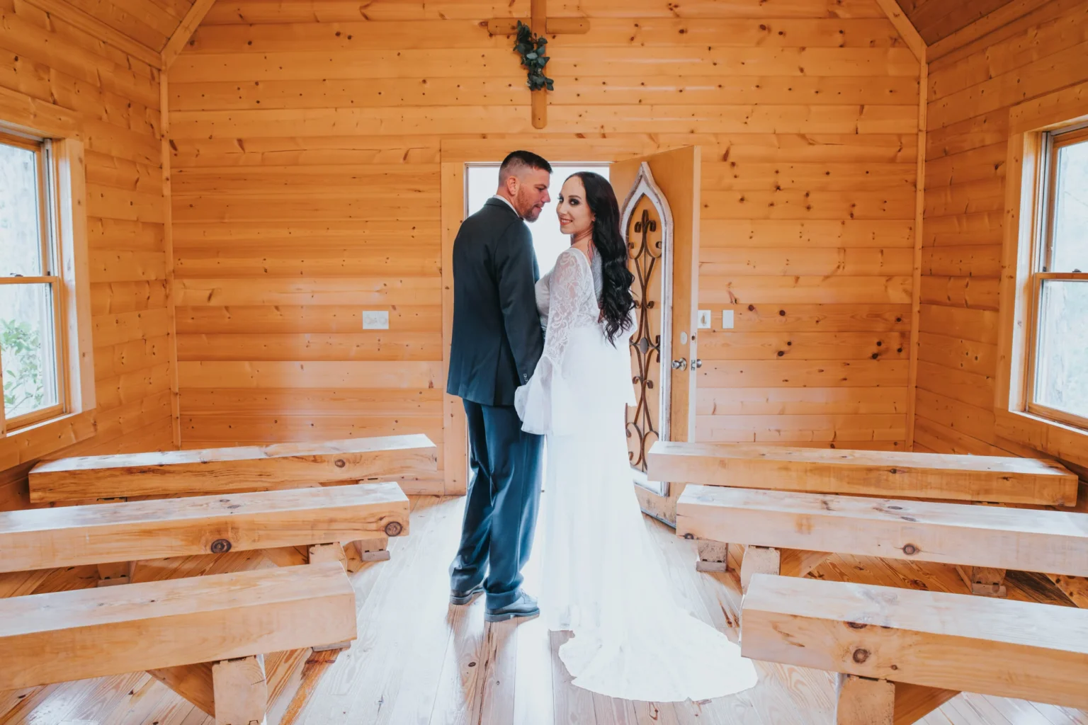 Elopement Wedding A couple stands in a small wooden chapel adorned with simple pews. The bride, in a white gown, and the groom, in a dark suit, share an intimate moment while holding hands and smiling at the camera. Sunlight streams in through the windows, creating a warm and inviting atmosphere. Elopements Inc