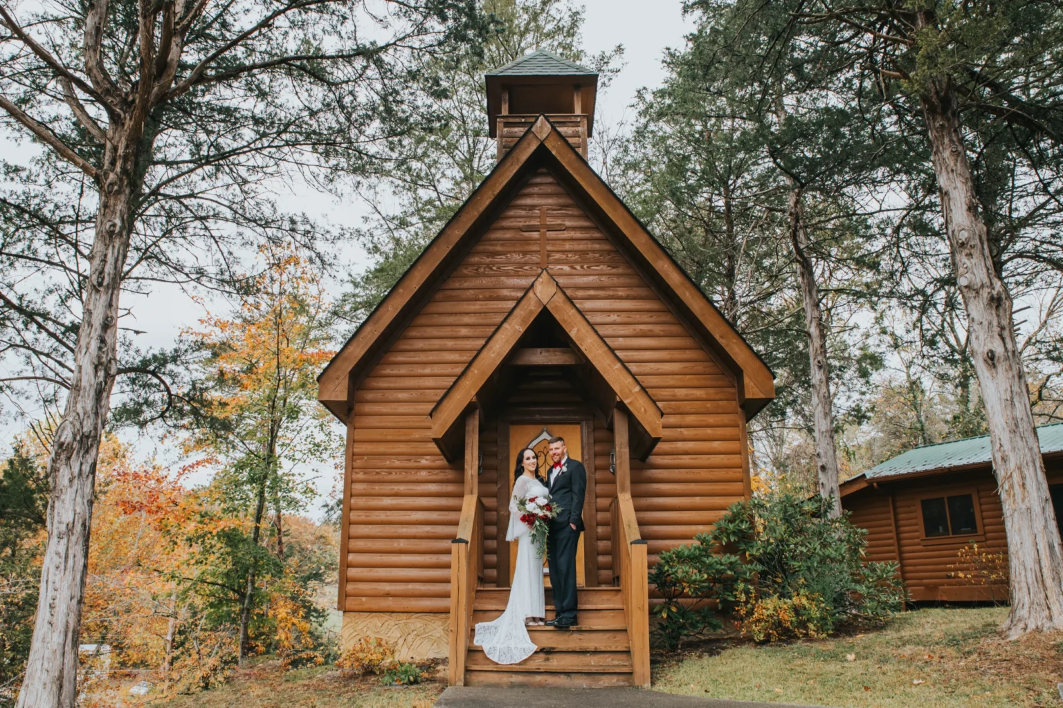 Elopement Wedding A bride and groom stand in front of a quaint wooden chapel surrounded by trees. The bride is in a white gown holding a bouquet, and the groom is in a suit. They are under the chapel's entrance awning, framed by the rustic structure and autumn foliage. The setting is serene and picturesque. Elopements Inc