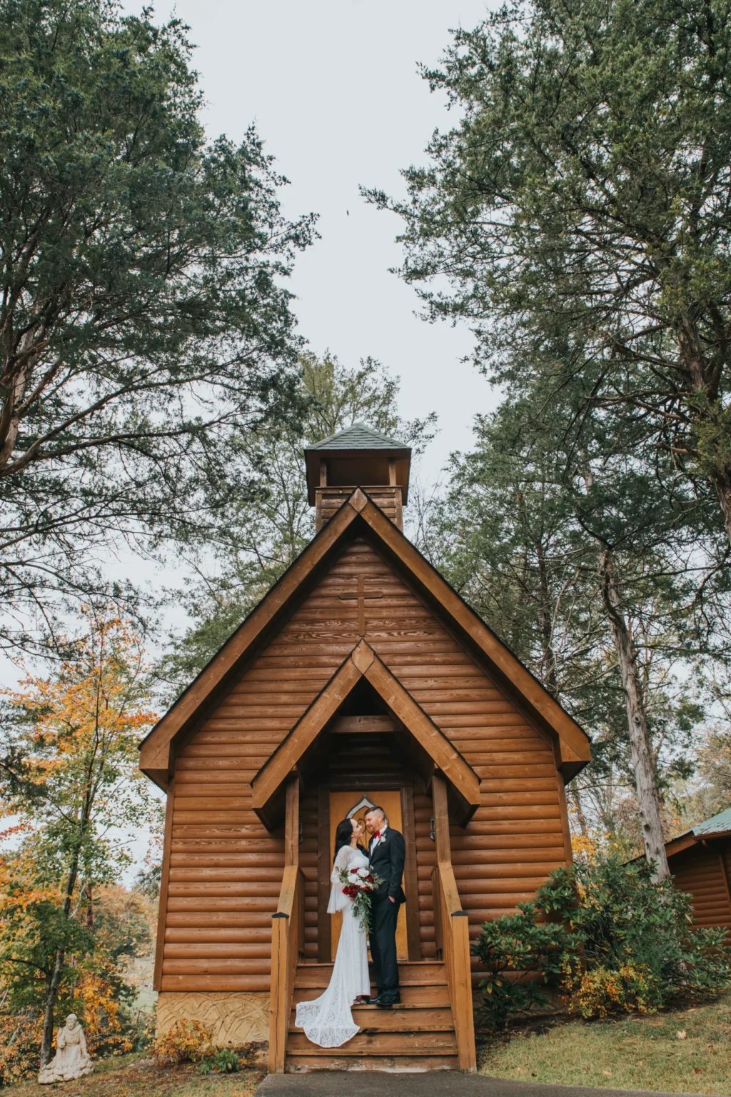 Elopement Wedding A couple stands on the steps of a quaint, small wooden chapel surrounded by tall trees. The bride, in a long white gown, holds a bouquet and faces her groom, who is dressed in a black suit. The chapel, made of dark logs with a steep roof, and the lush greenery create a serene and romantic atmosphere. Elopements Inc