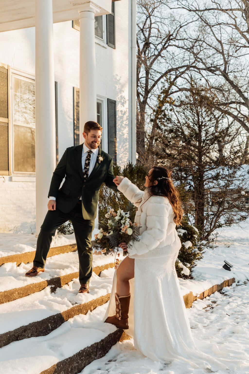 Elopement Wedding A couple stands on snowy steps in front of a white building with columns. The groom, in a dark green suit and patterned tie, holds the hand of the bride, who is in a white dress, white jacket, and boots, carrying a bouquet. Bare trees and sunlight in the background suggest a winter wedding. Elopements Inc