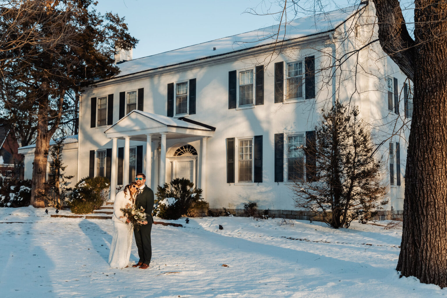 Elopement Wedding A couple stands in front of a large white house with black shutters and a columned portico. The ground is covered with snow and the trees are bare. The couple is in wedding attire, with the bride in a white dress and veil, and the groom in a dark suit with a tie. Both are smiling and embracing. Elopements Inc