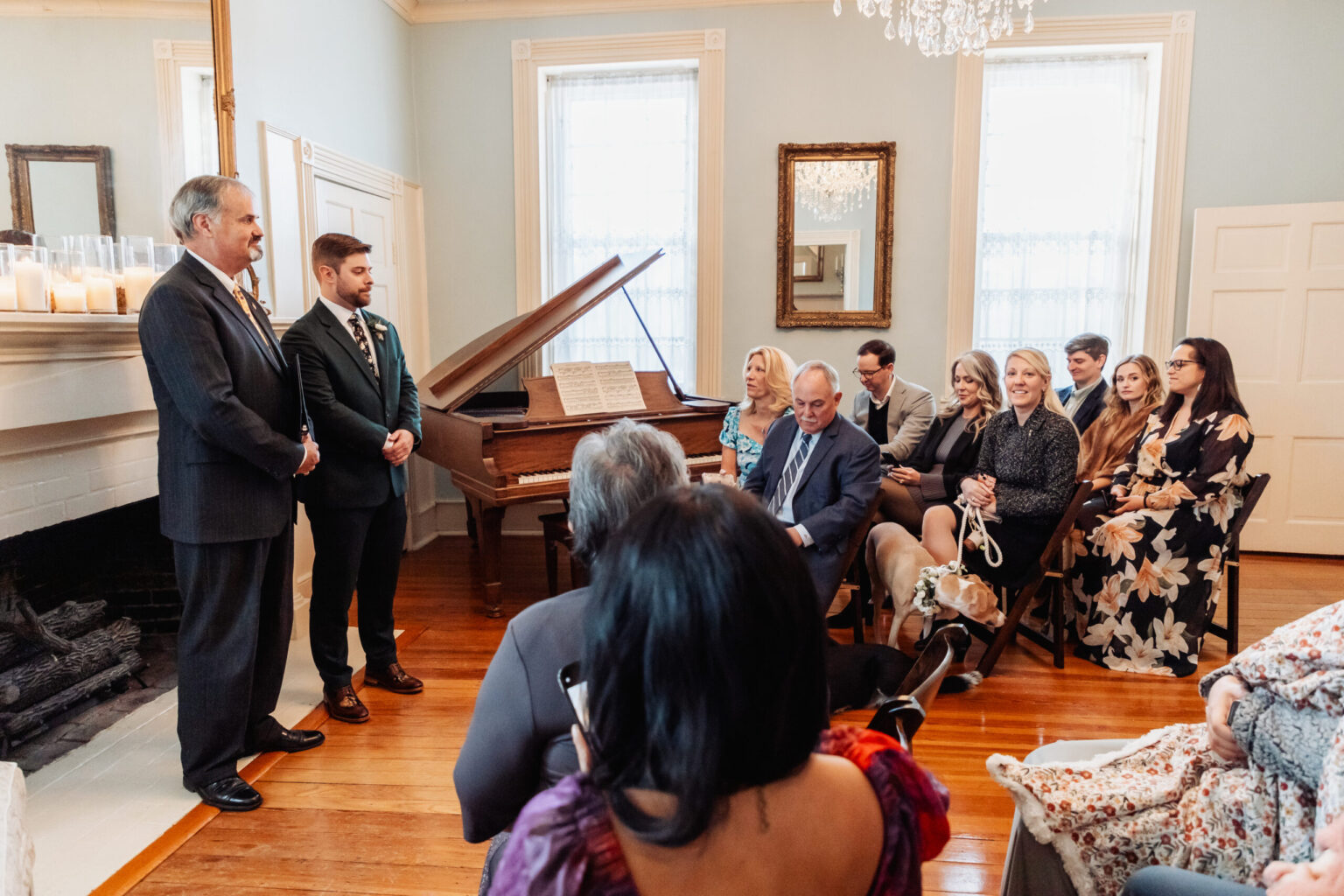 Elopement Wedding A room with people gathered for a formal event. Two men in suits stand beside a grand piano, with one looking at guests who are seated facing them. Attendees are dressed in formal attire, and a chandelier hangs from the ceiling. The room has large windows, mirrors, and a fireplace with candles. Elopements Inc