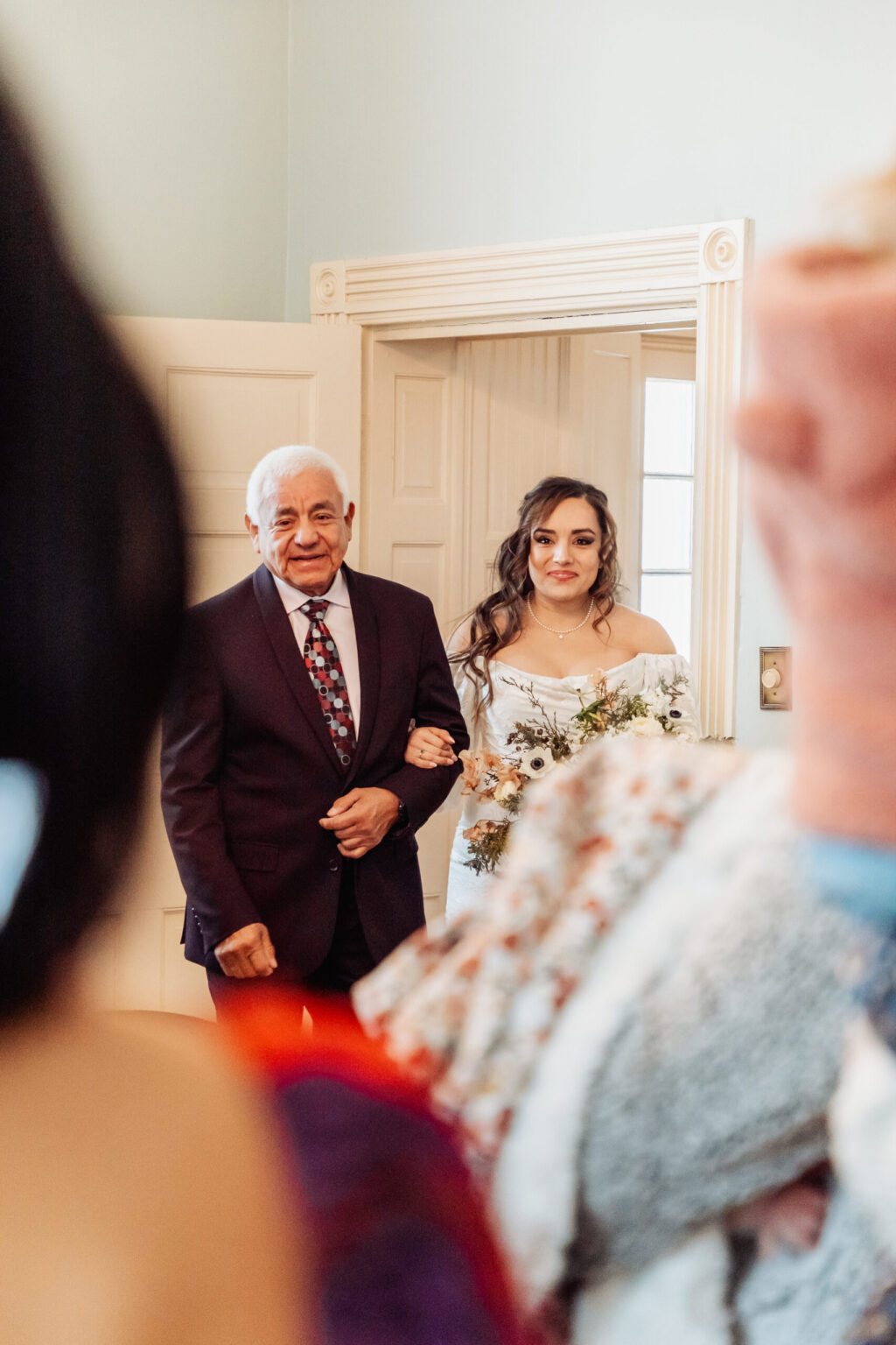 Elopement Wedding A bride holding a bouquet of flowers walks arm-in-arm with an elderly man through a doorway. She wears an off-shoulder white wedding gown and has long, wavy hair. The man wears a dark suit, white shirt, and patterned tie. They both look forward and smile. Other people are visible in blurred foreground. Elopements Inc