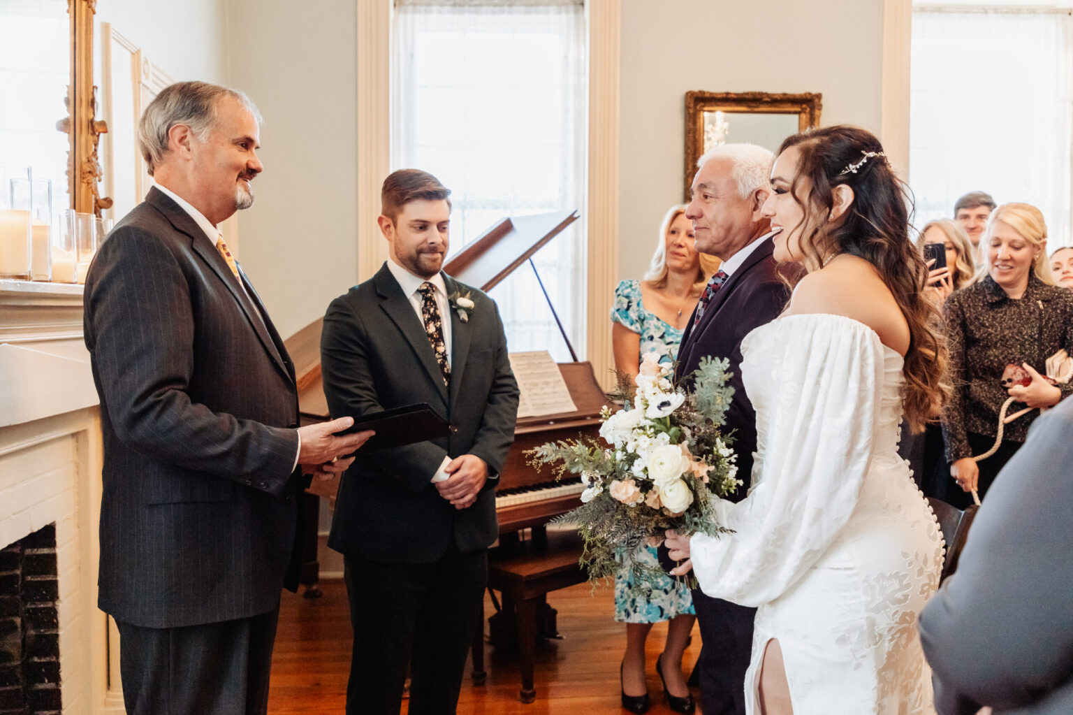 Elopement Wedding A bride in a white off-shoulder gown stands beside an older man in a suit at a wedding ceremony. They face an officiant holding a book, with a groom in a dark green suit standing beside him. Guests watch from the background in a room with tall windows and a piano. The bride holds a bouquet. Elopements Inc