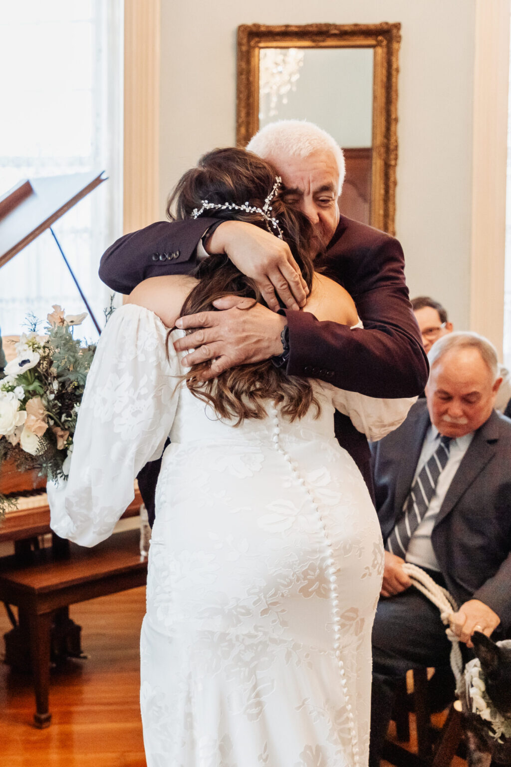 Elopement Wedding A bride in a white, long-sleeved wedding dress with lace detail hugs an older man in a dark suit. The bride's hair is styled with a decorative headpiece. They are standing in a warmly lit room with people seated and a large mirror on the wall behind them. A flower arrangement is visible to the left. Elopements Inc