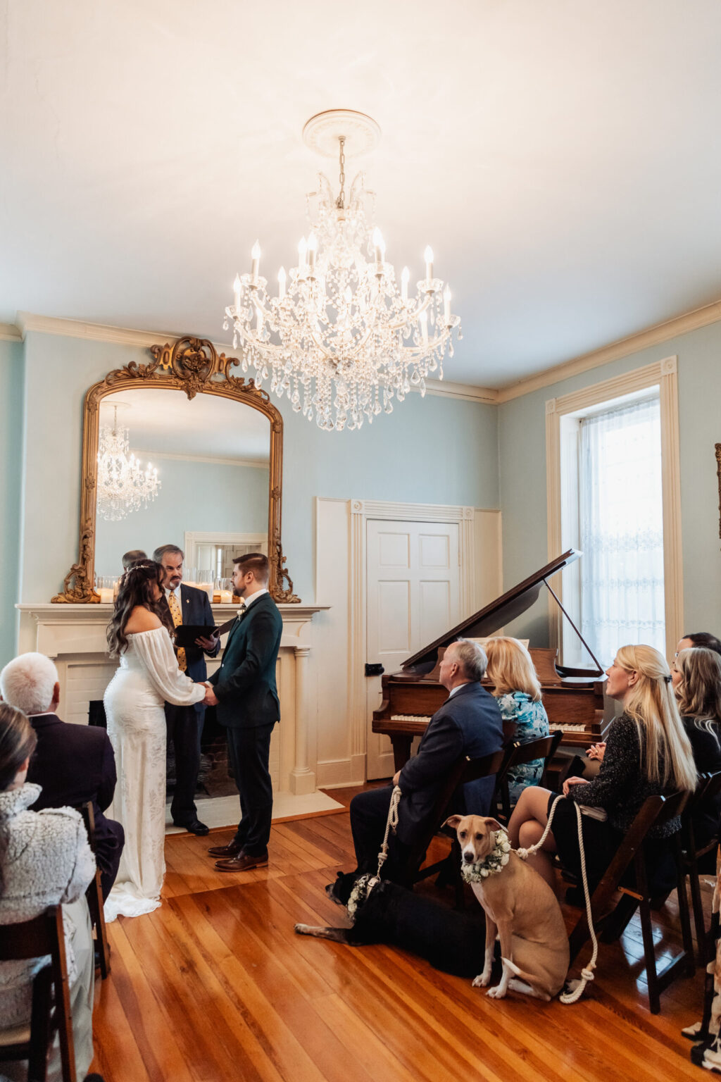 Elopement Wedding A bride and groom stand facing each other holding hands during a wedding ceremony in a elegant room with hardwood floors. An officiant stands between them. Seated guests, including a dog on a leash, watch attentively. A grand piano and ornate chandelier adorn the space. Elopements Inc