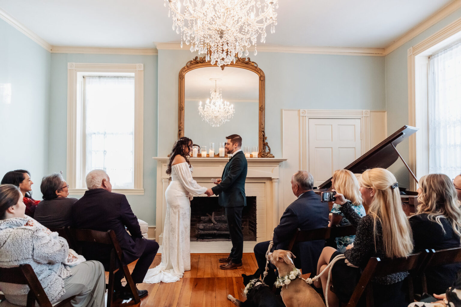 Elopement Wedding A bride and groom stand facing each other in a small, elegant room, exchanging vows. Guests are seated on dark wooden chairs, watching attentively. The room features a chandelier, a large mirror above a fireplace, and a piano on the right. Two dogs are lying down near the guests. Elopements Inc