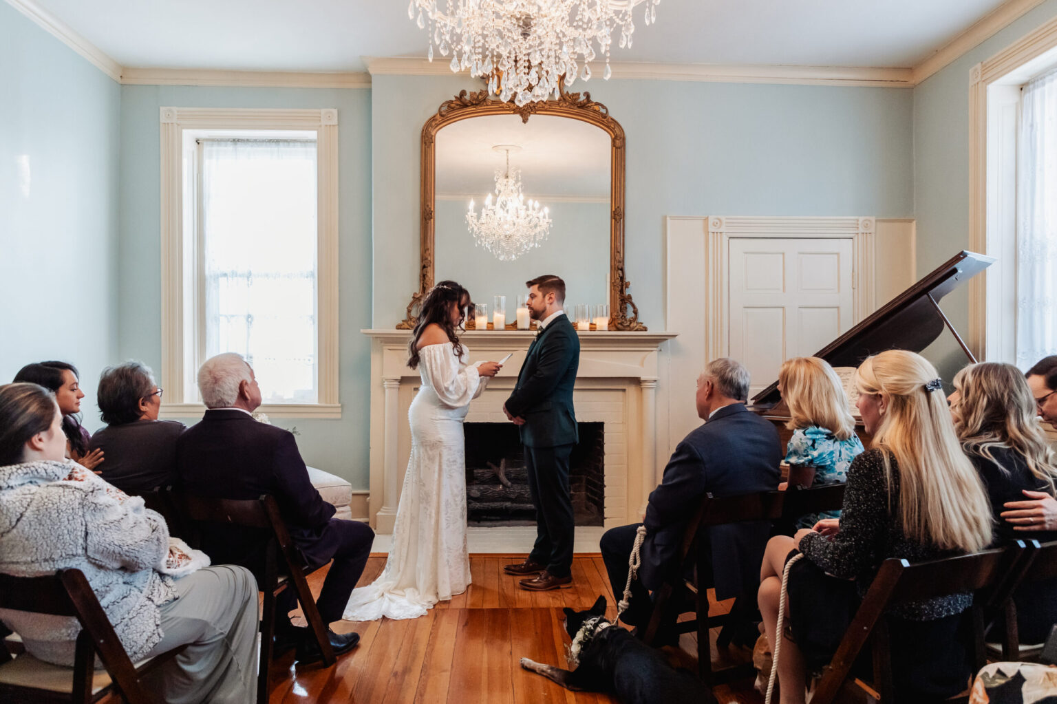 Elopement Wedding A bride and groom stand in front of a fireplace exchanging vows in an elegantly decorated room with pale blue walls. Guests are seated on dark wooden chairs. A chandelier hangs from the ceiling, and there is a grand piano by the window. A dog lies on the floor near the guests. Elopements Inc